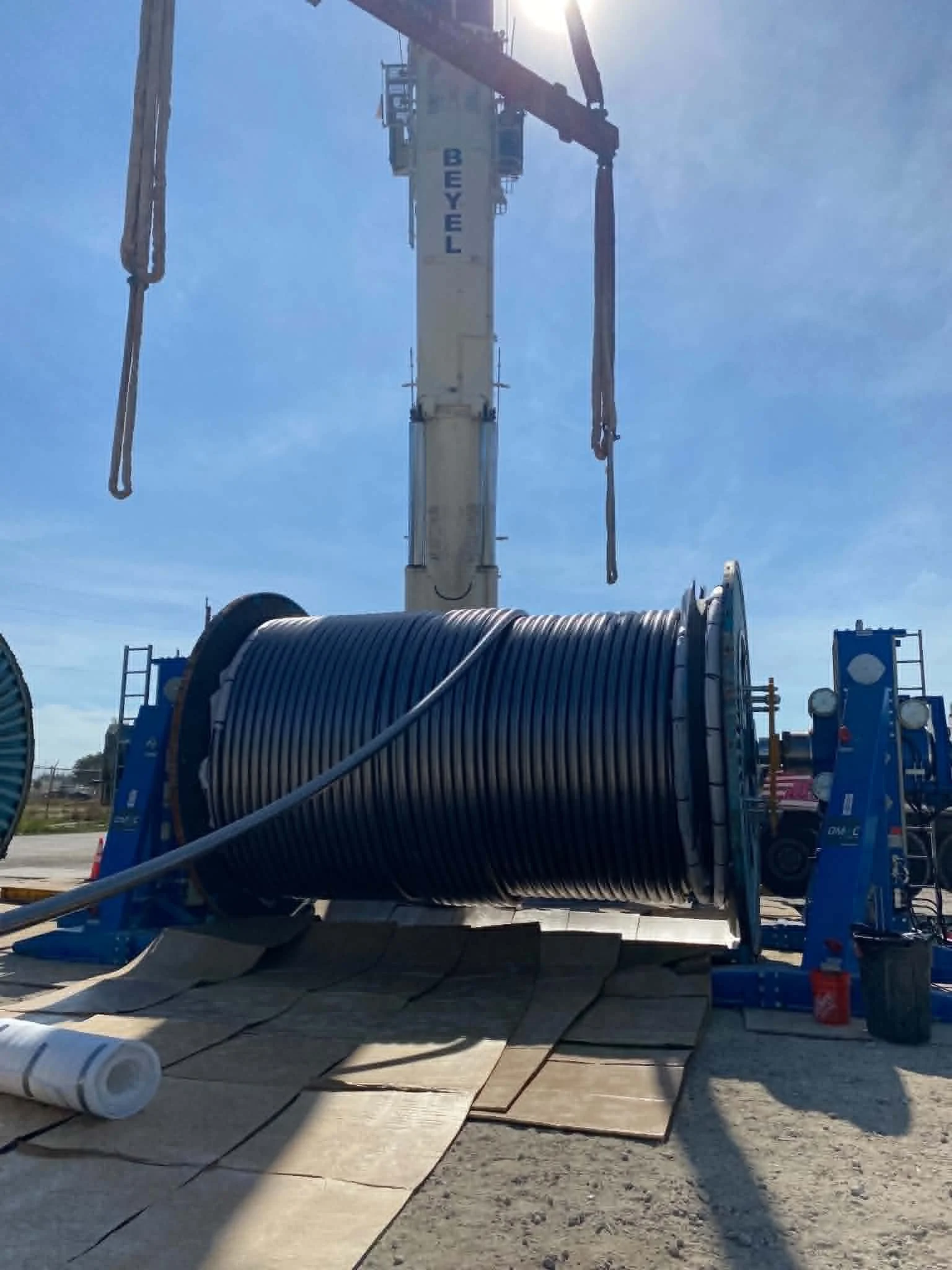 Construction site with a large crane lifting the arm and pipes, with a conveyor system, large rolled pipe, and wooden planks on the ground, under clear blue sky.