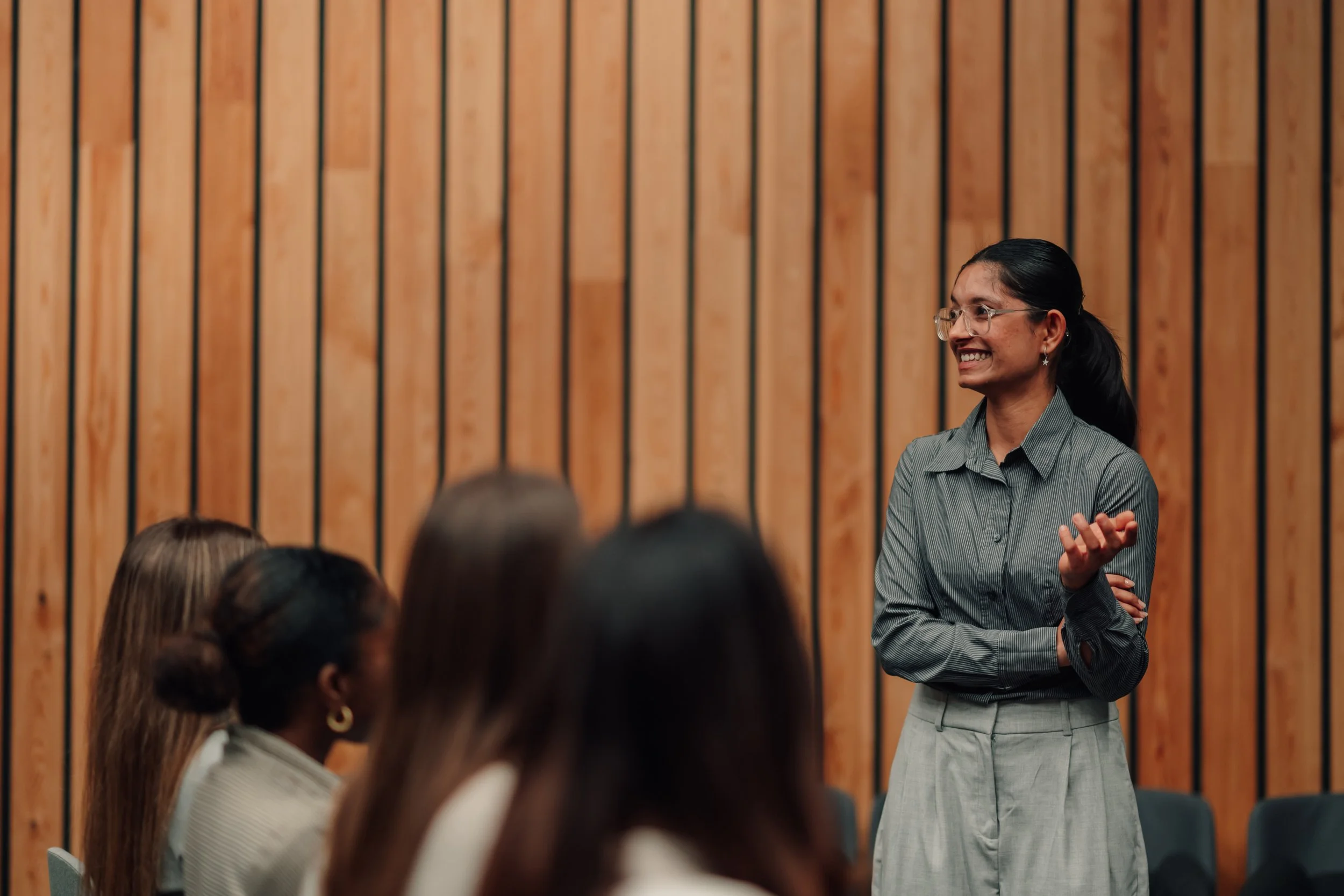 A woman with glasses, dark hair in a ponytail, wearing a striped shirt, smiling and speaking to a group in a room with wooden panel walls.
