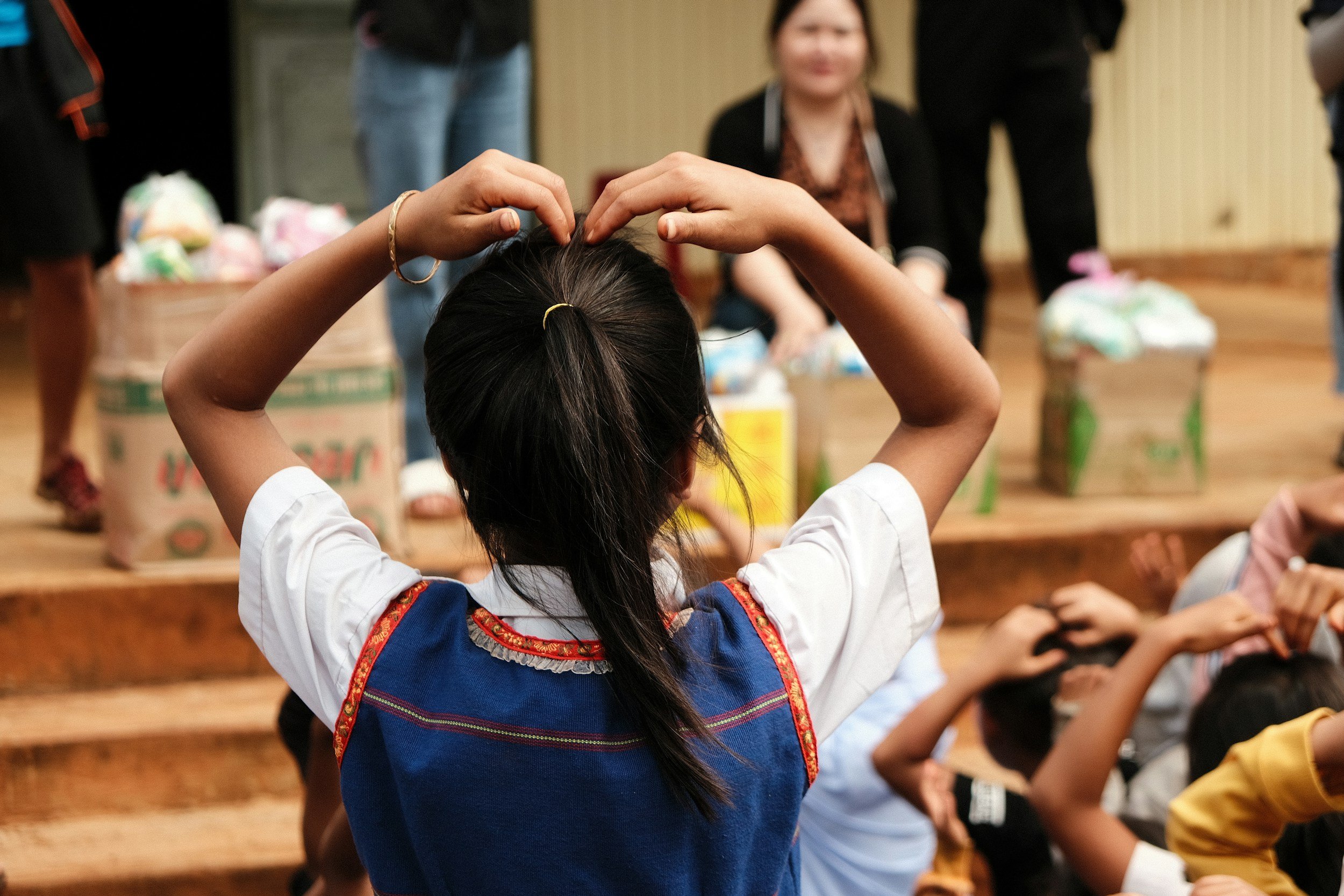A child with long dark hair, wearing a traditional dress with-colored embroidery, forms a heart shape above her head with her hands. Several other children and adults are visible in the background, sitting at tables with food or supplies in a warm indoor setting.