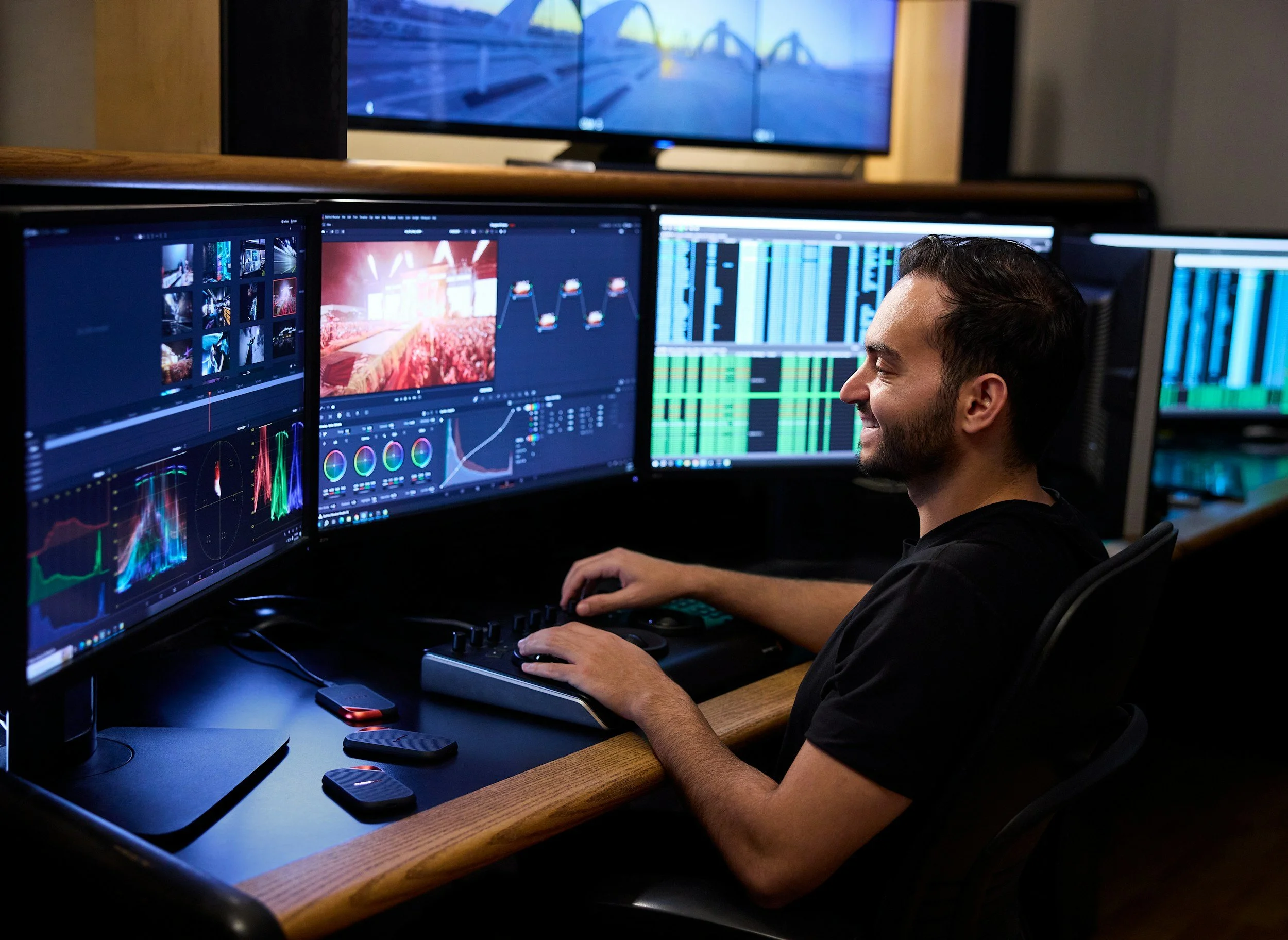 A man working at a desk with three computer monitors displaying video editing and audio editing software, in a dimly lit room with a large TV screen mounted above displaying a bridge scene.