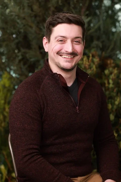 A smiling man sitting outdoors with trees in the background.