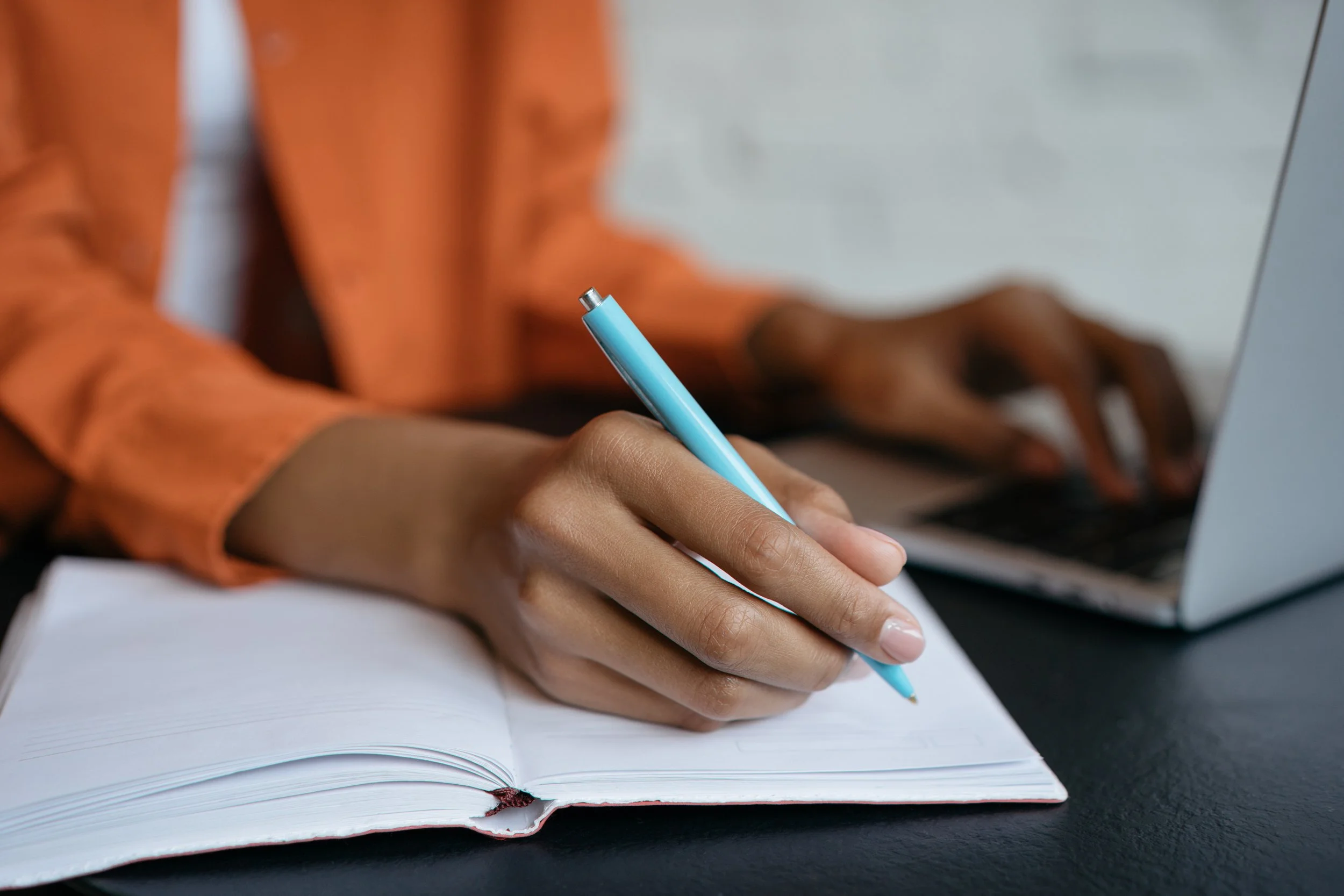 Close-up of a hand holding a blue pen, writing in a white notebook on a dark surface. The person is wearing an orange long-sleeve shirt.