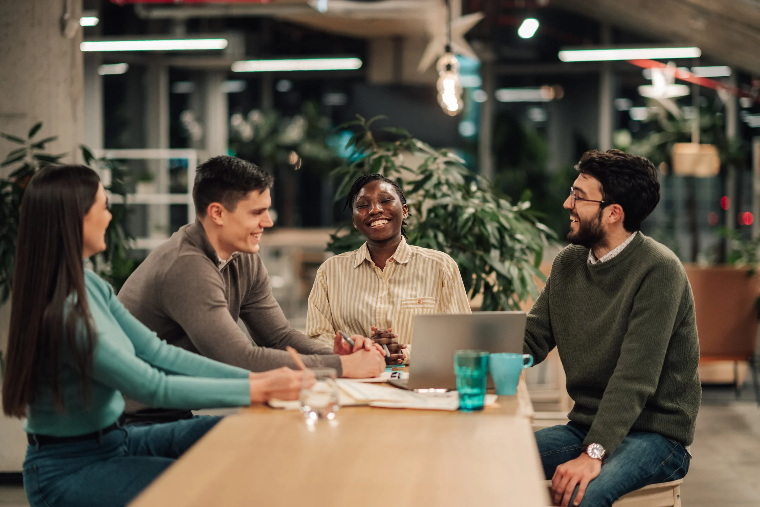 Group of five diverse young adults smiling and engaging in conversation around a table in a modern office or coworking space, with plants and hanging lights in the background.