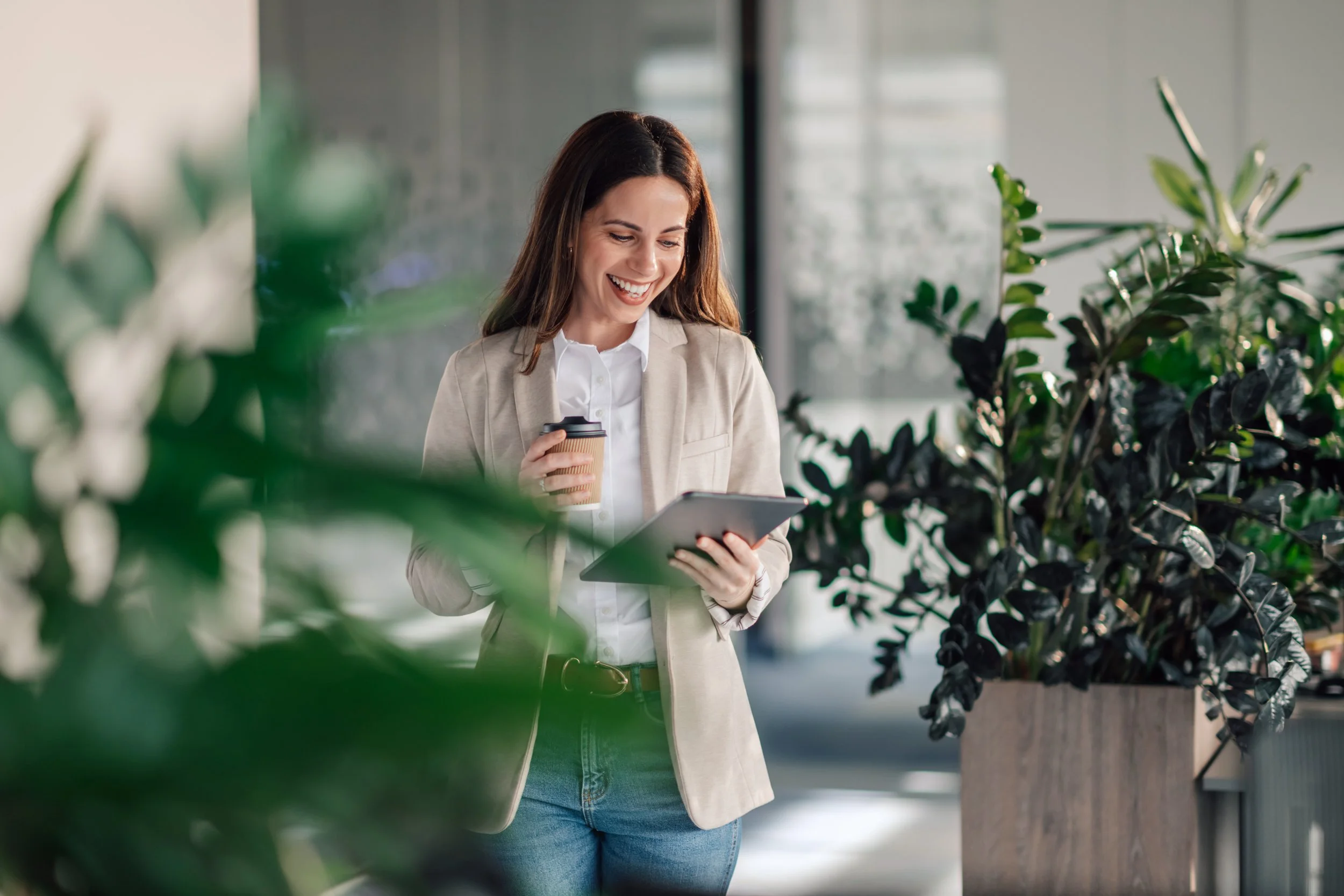 A woman in a beige blazer and white shirt, holding a disposable coffee cup and a tablet, smiling while looking at the tablet in a modern indoor setting with green plants.