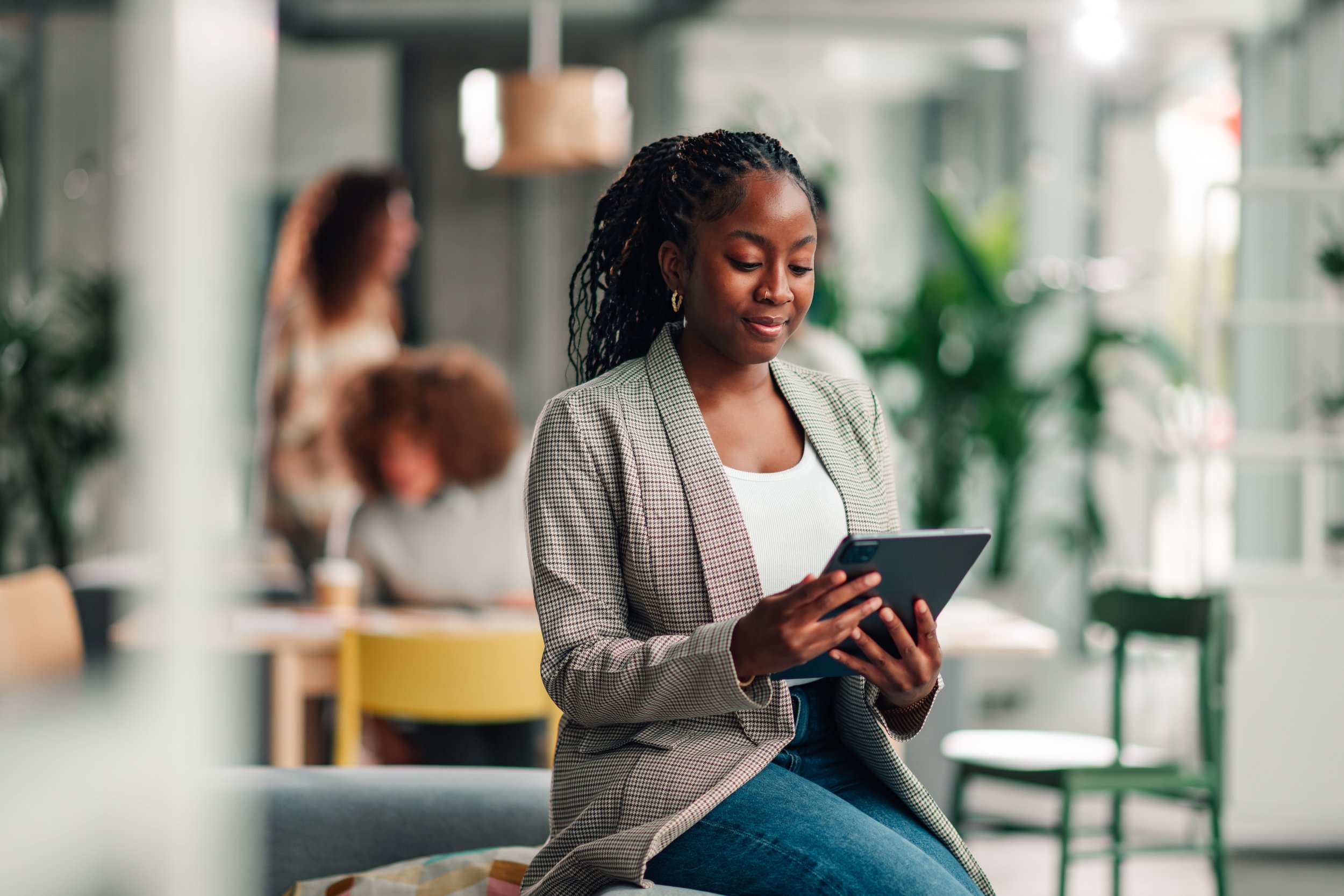 A woman with braids and a checkered blazer sitting on a bench, looking at her smartphone, in a bright indoor space with green plants and blurred people in the background.