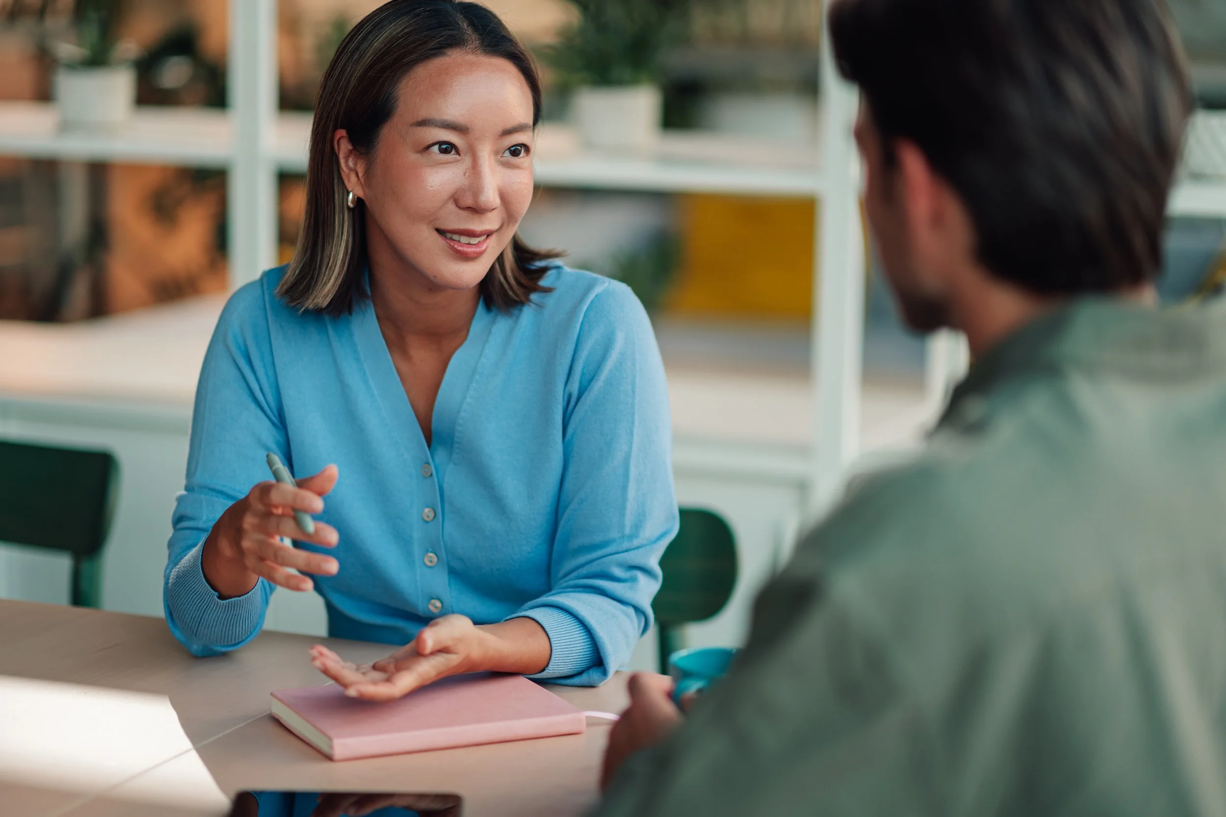 A woman with shoulder-length hair, wearing a light blue sweater, engaged in conversation with a man. She is gesturing with her left hand and holding a pen in her right hand. They are sitting at a table with a pink notebook. The background shows a shelf with plants and decorative items.