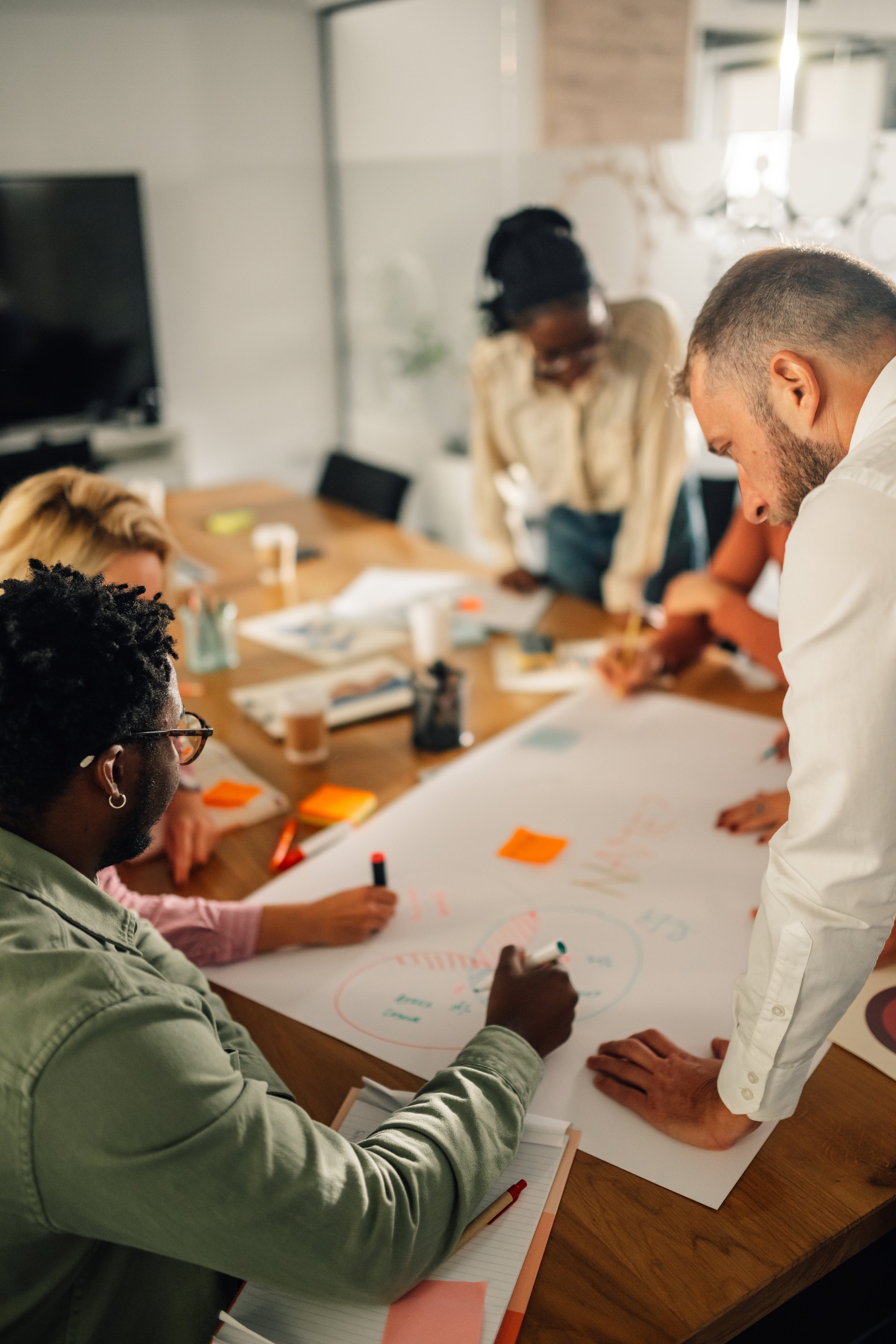 A group of diverse people working together around a table with large paper, markers, and notebooks, collaborating on a project.