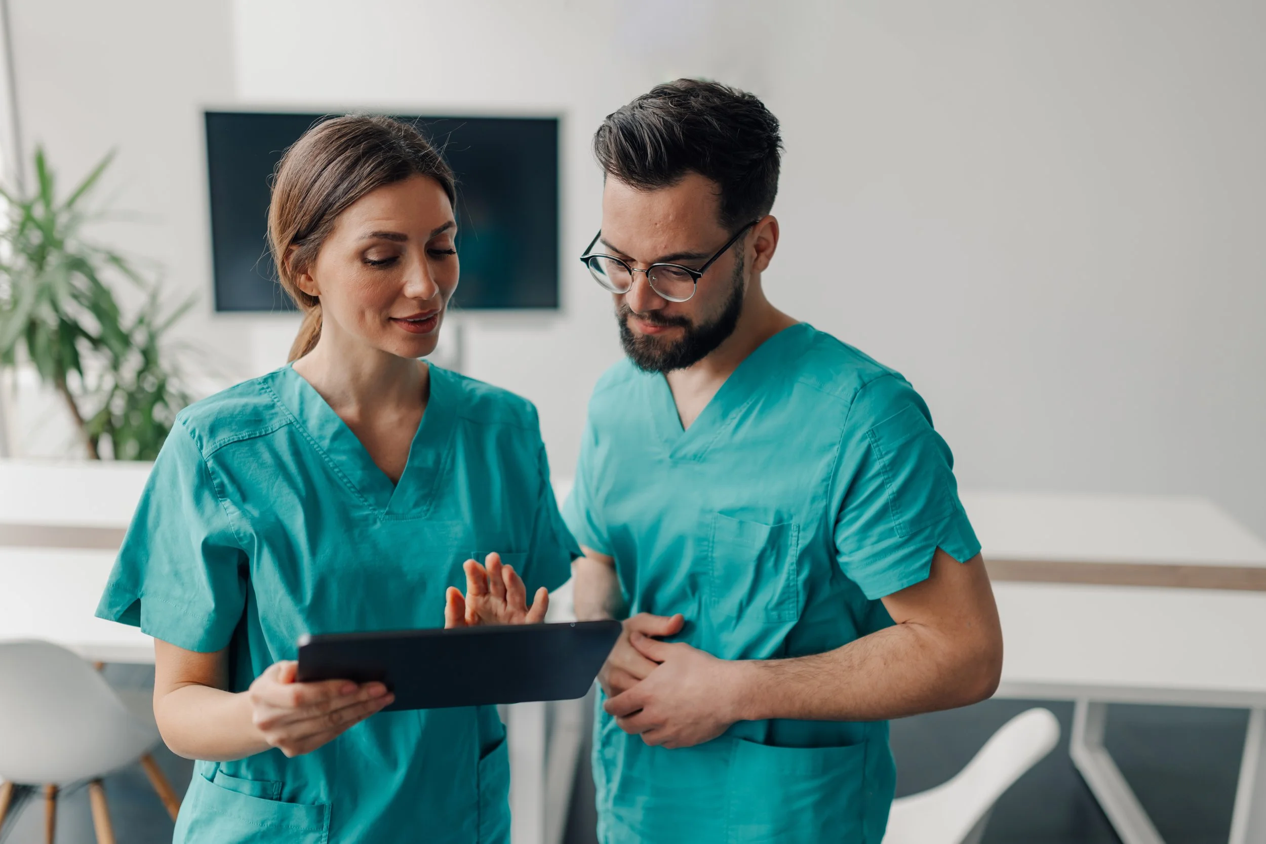 Two healthcare professionals, a woman and a man, wearing blue scrubs, looking at a tablet together in a bright, modern medical office.