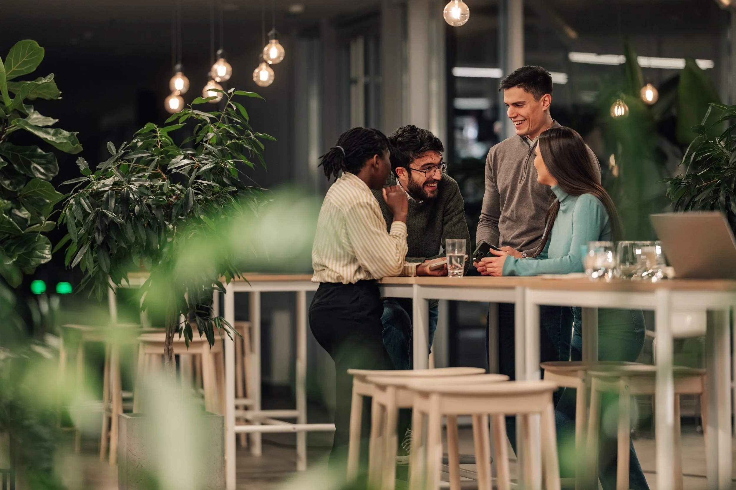Four young adults are gathered around a table, smiling and engaged in conversation inside a modern, well-lit café or restaurant with hanging light bulbs and large green plants.