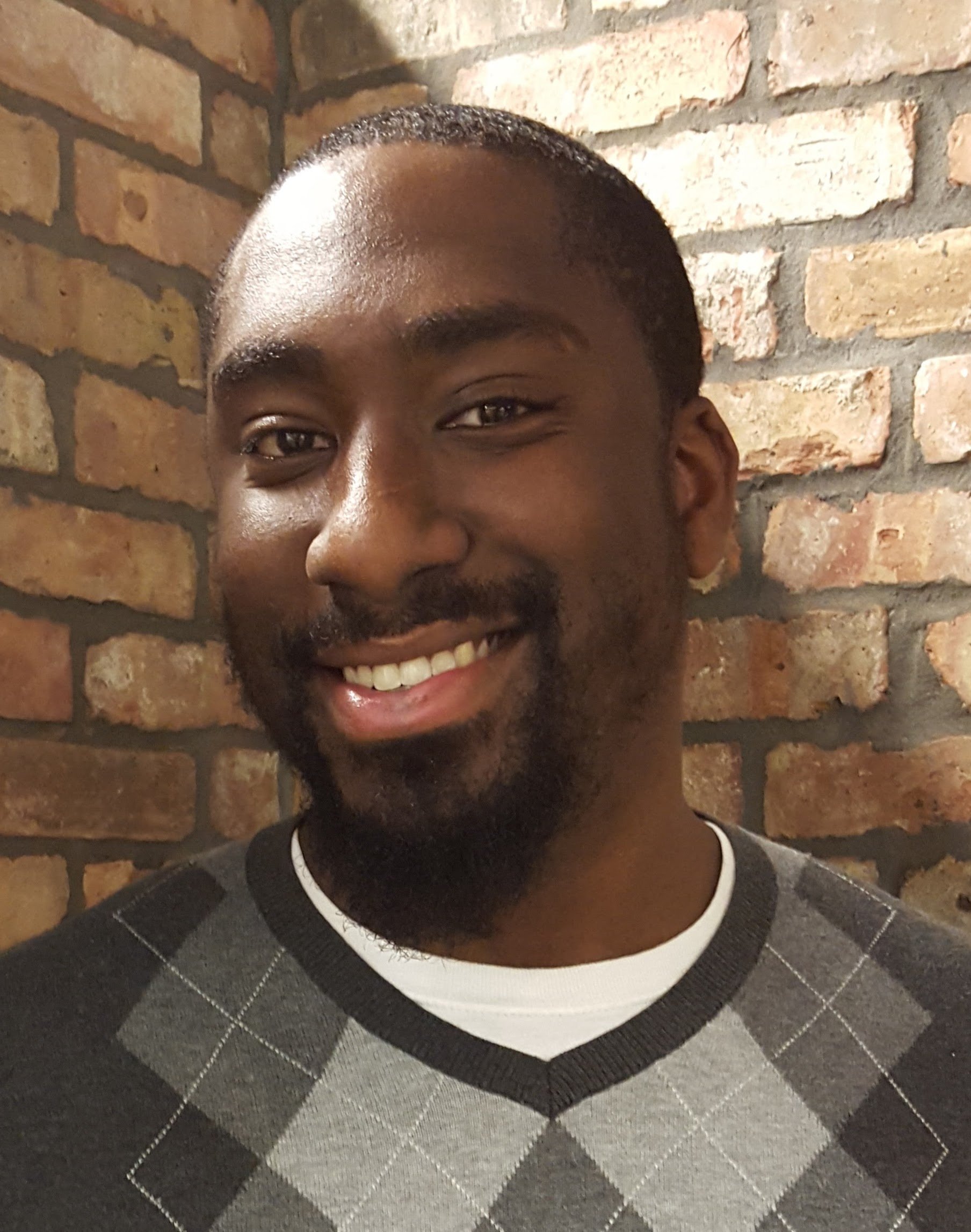 Close-up of a smiling man's face against a brick wall, showing his eyes, nose, and mouth, with short hair and facial hair, wearing a patterned sweater.