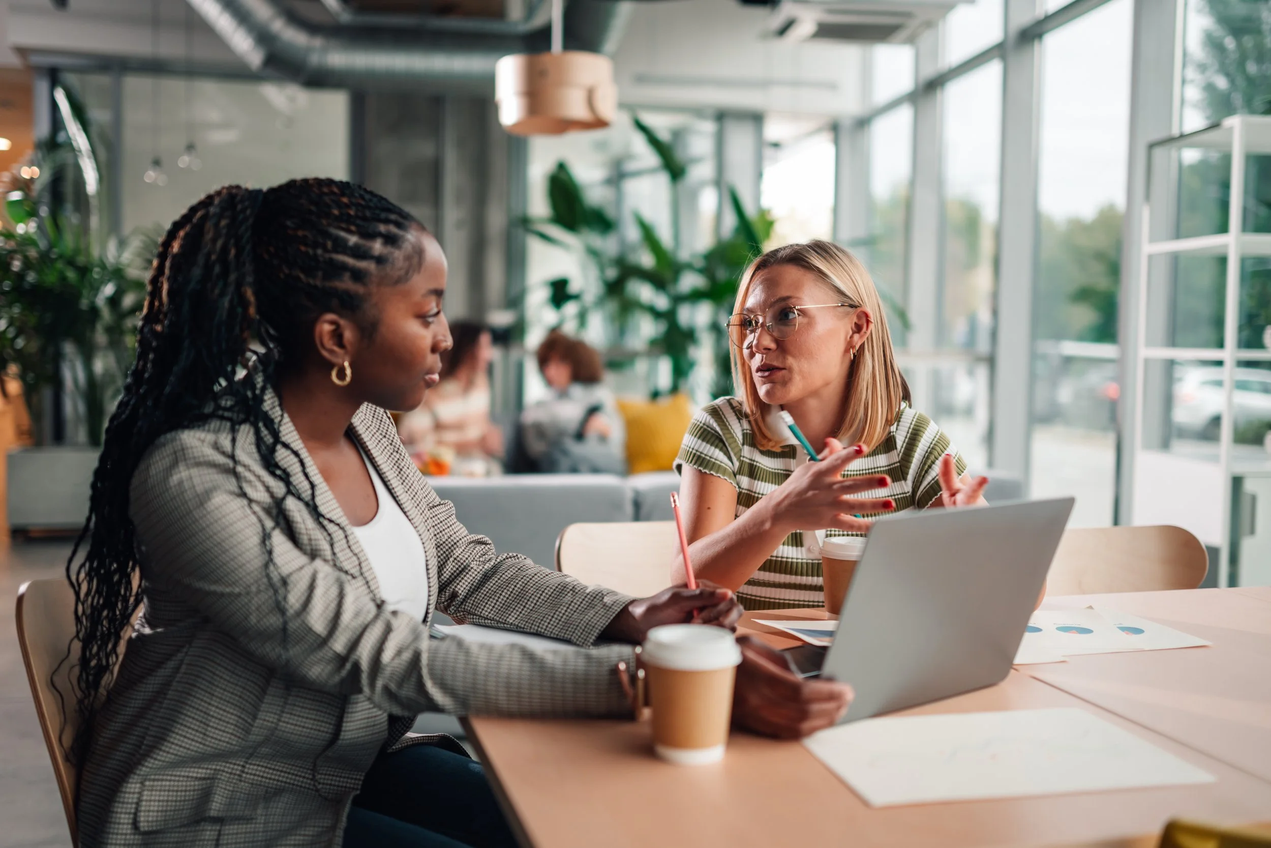 Two women having a conversation at a table in a bright, modern cafe with large windows and green plants. One woman has dark hair with braids, and the other has blonde hair with glasses. There are cups, a laptop, and papers on the table.
