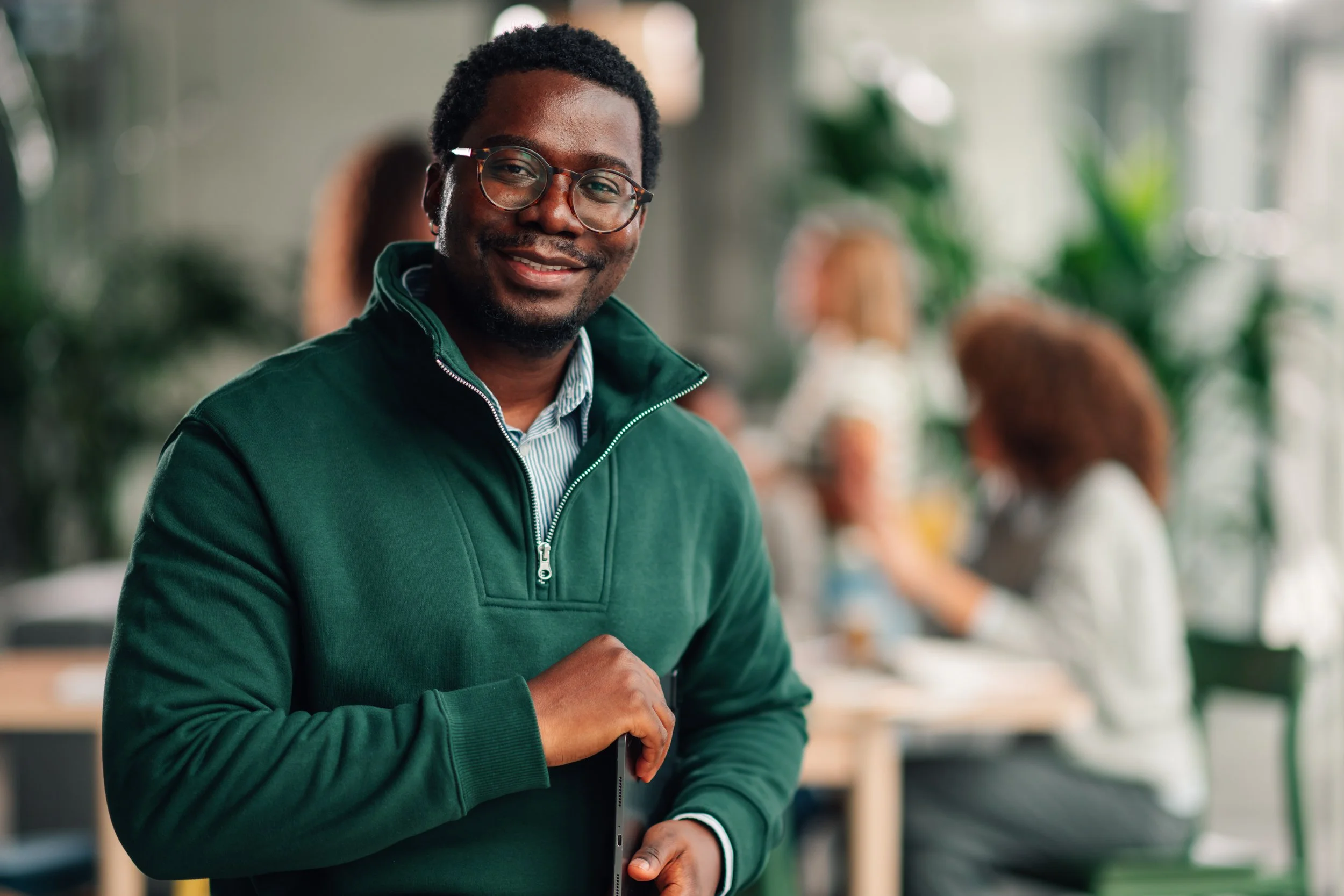 A man with glasses and a dark green jacket smiling inside a modern, well-lit space with blurred background of people and greenery.