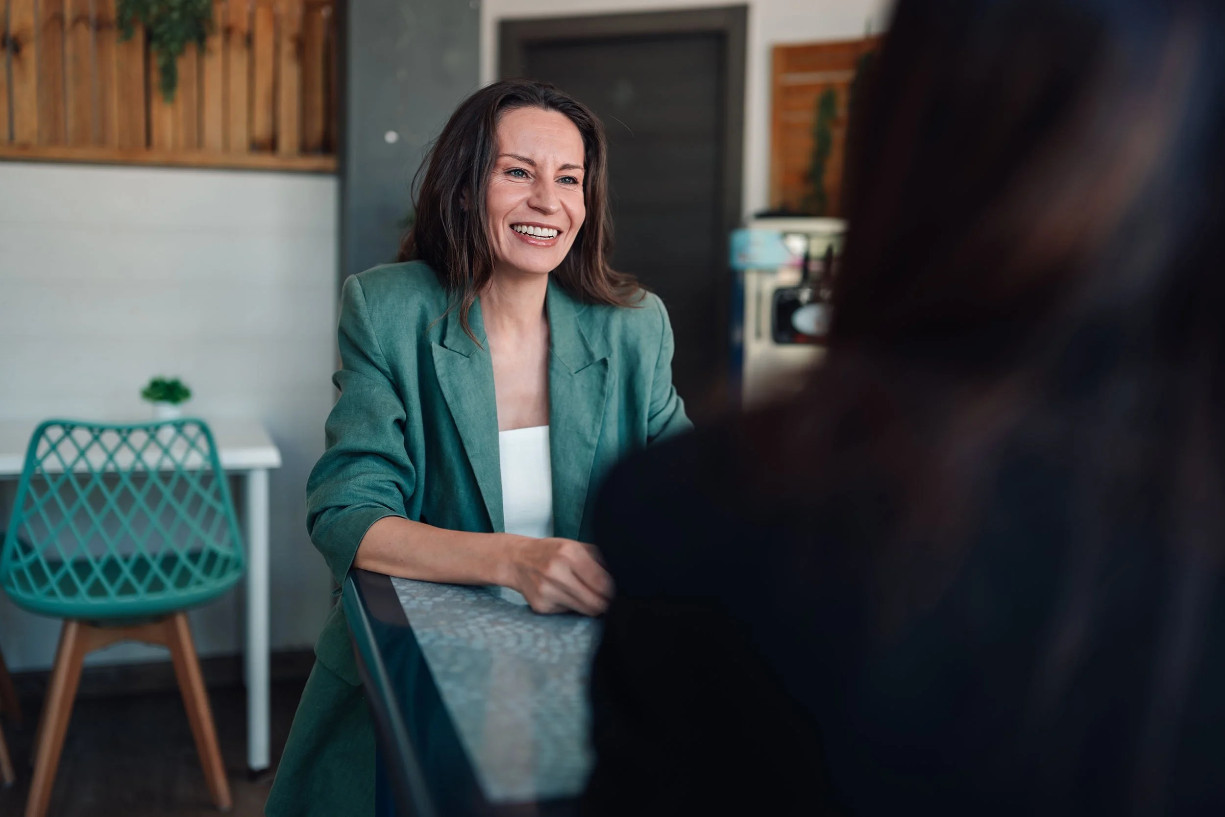 A woman smiling and talking to a person sitting at a table in a cafe or restaurant.