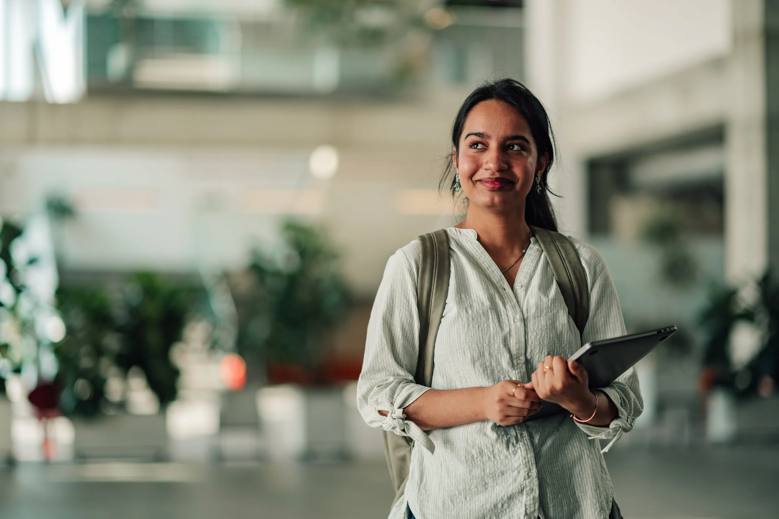 Young woman with dark hair and earrings, smiling, holding a tablet, in a modern indoor space with blurred background of plants and architecture.
