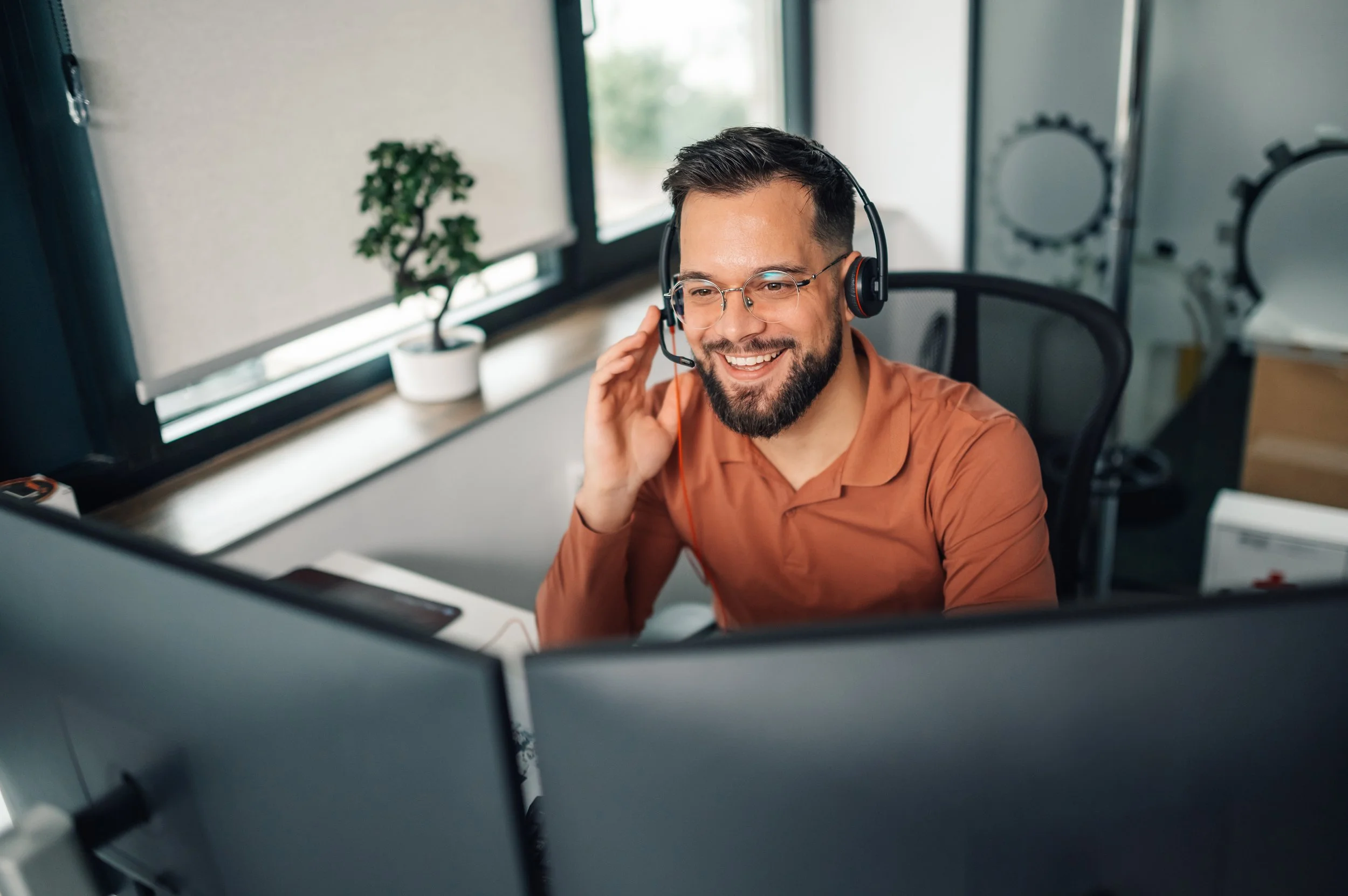 A man with glasses and a beard wearing an orange shirt, smiling, and wearing a headset while working at his desk in an office. There are windows with blinds, a small potted plant, and other office equipment visible in the background.