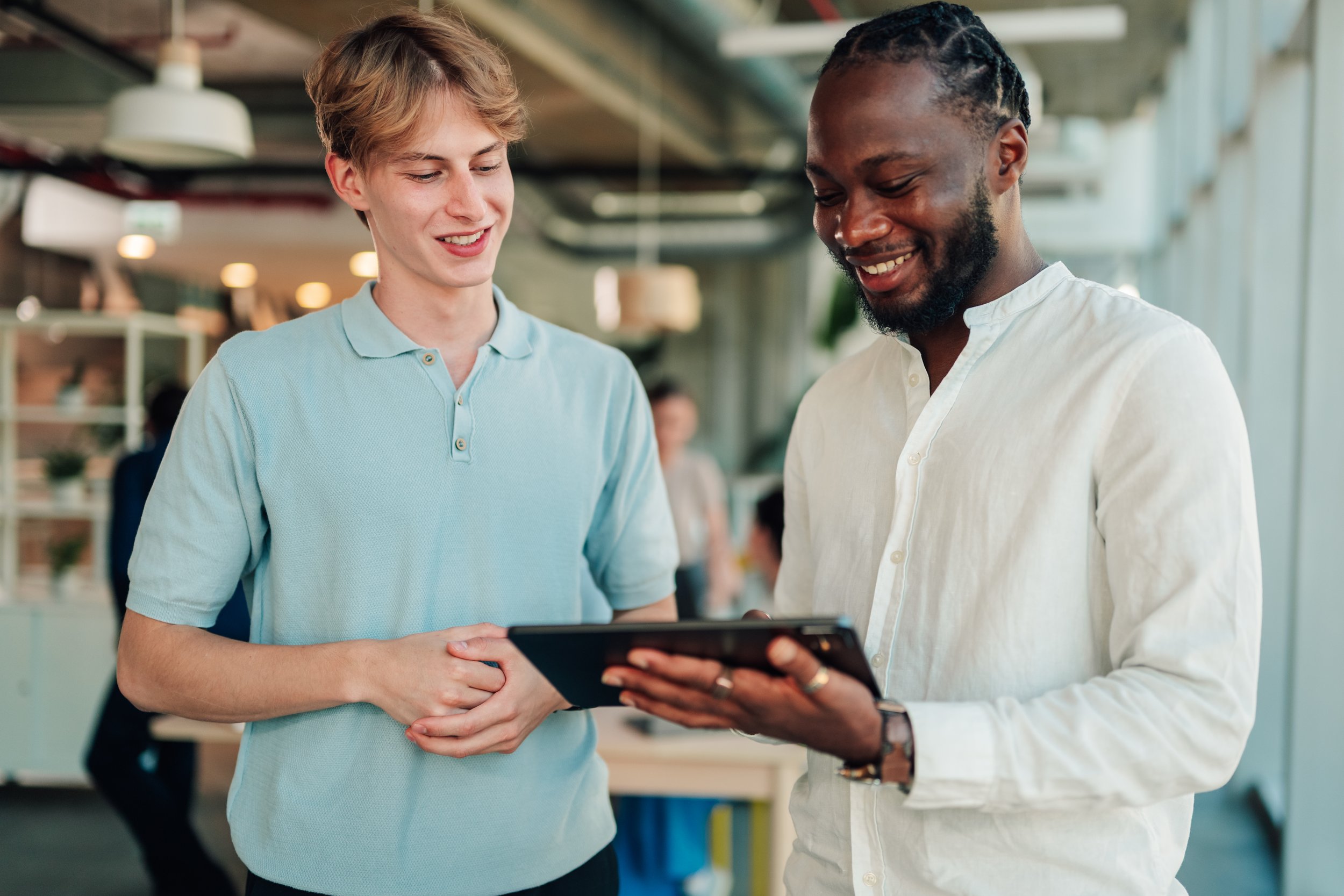 Two smiling men in a modern office, looking at a tablet device together.