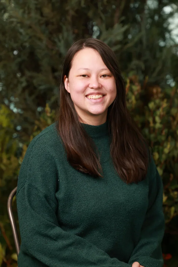 A young woman with long brown hair and a broad smile, wearing a dark green sweater, posing outdoors with blurred green foliage in the background.