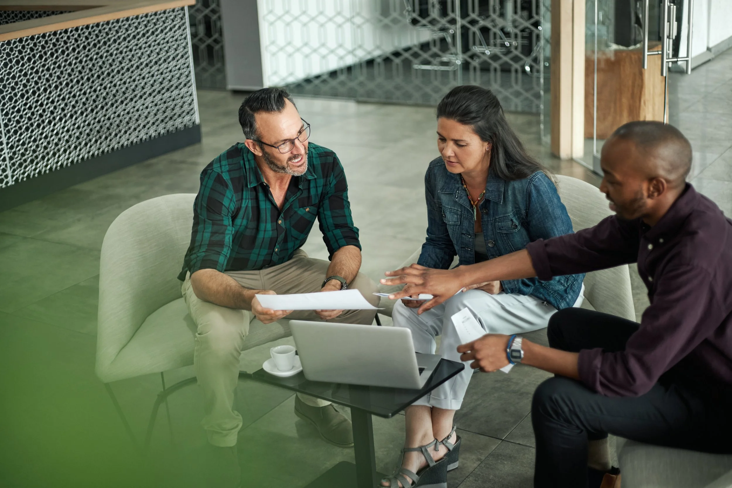 Three people sitting around a table in a modern office space, discussing documents and working on laptops.