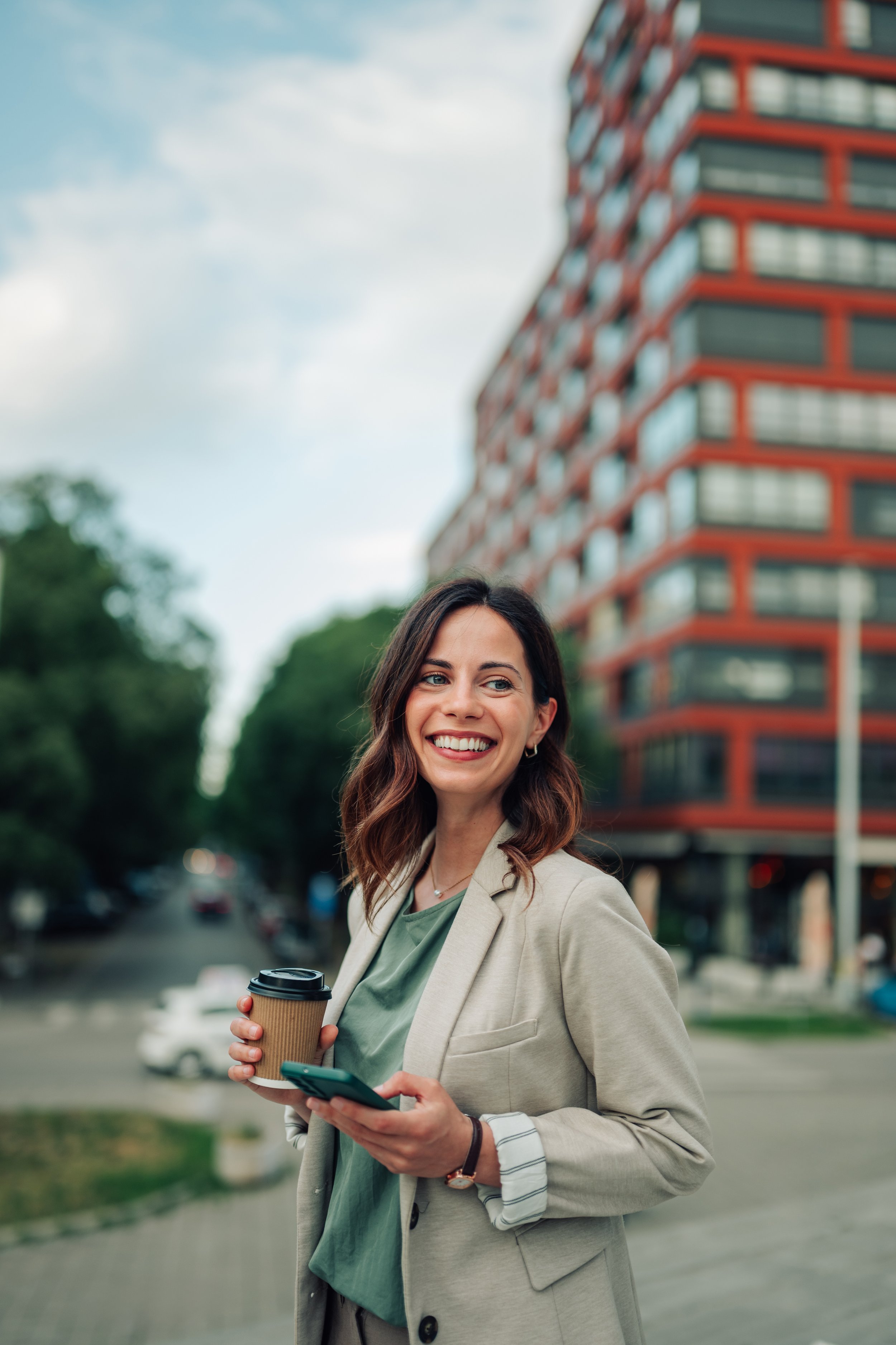 A woman with shoulder-length brown hair, wearing a beige blazer and a green top, smiling and holding a coffee cup and a smartphone while standing outdoors on a city street with a modern red building and trees in the background.