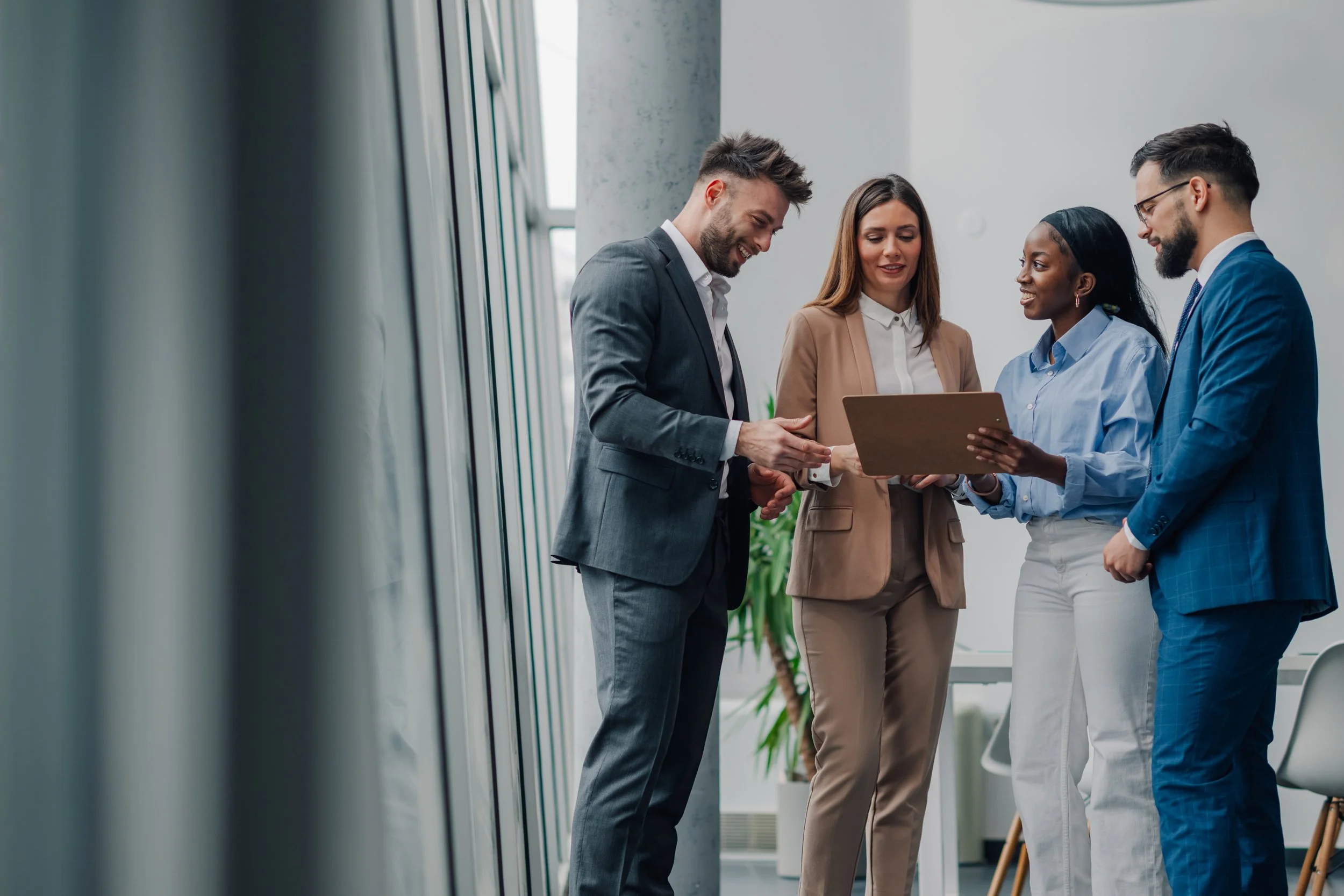 Four diverse professionals in business attire engaged in conversation in a modern office corridor, with large windows and greenery.