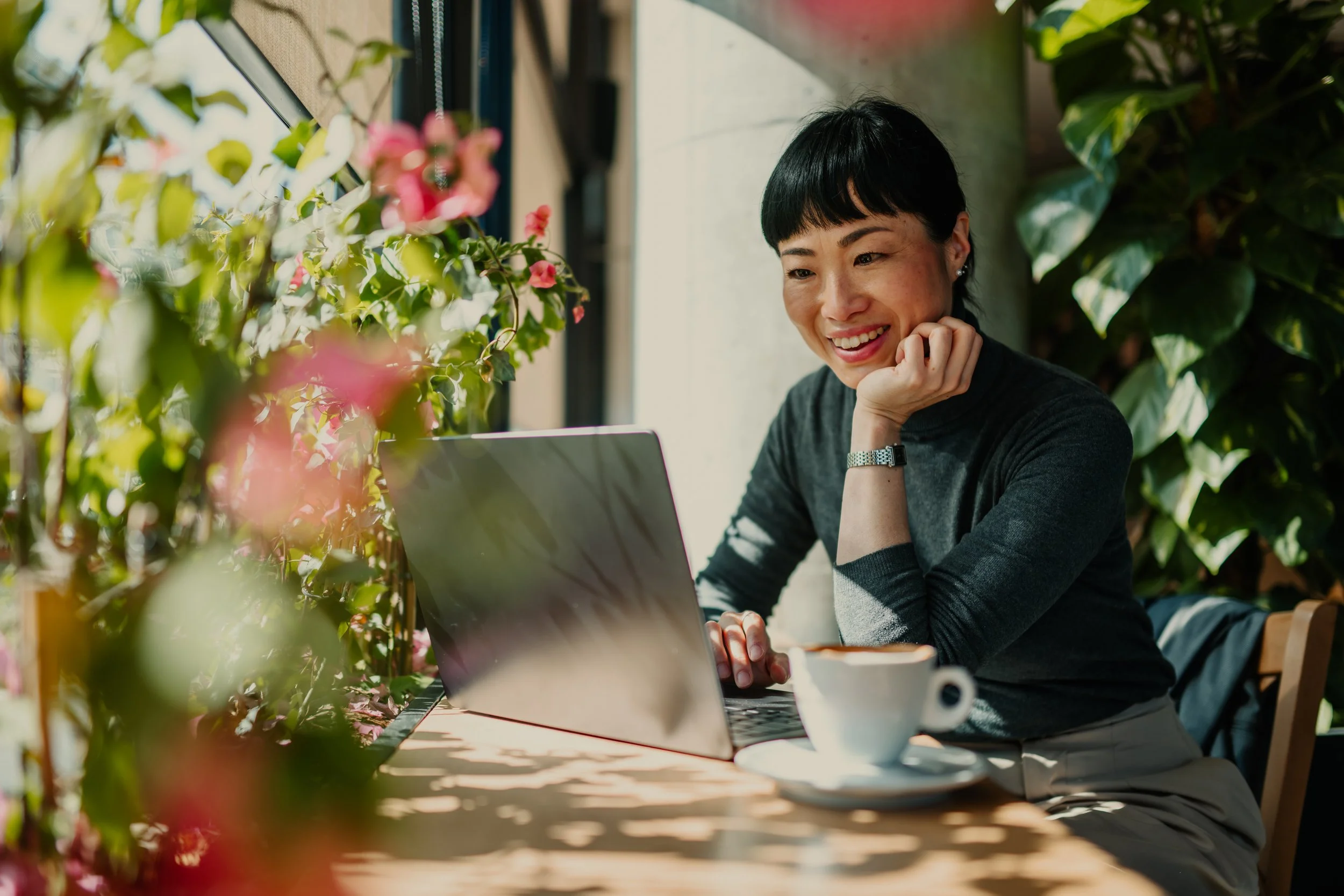 A woman with short black hair and a black and gray sweater sitting at a table with a laptop, smiling while looking at the screen, surrounded by green plants and pink flowers.