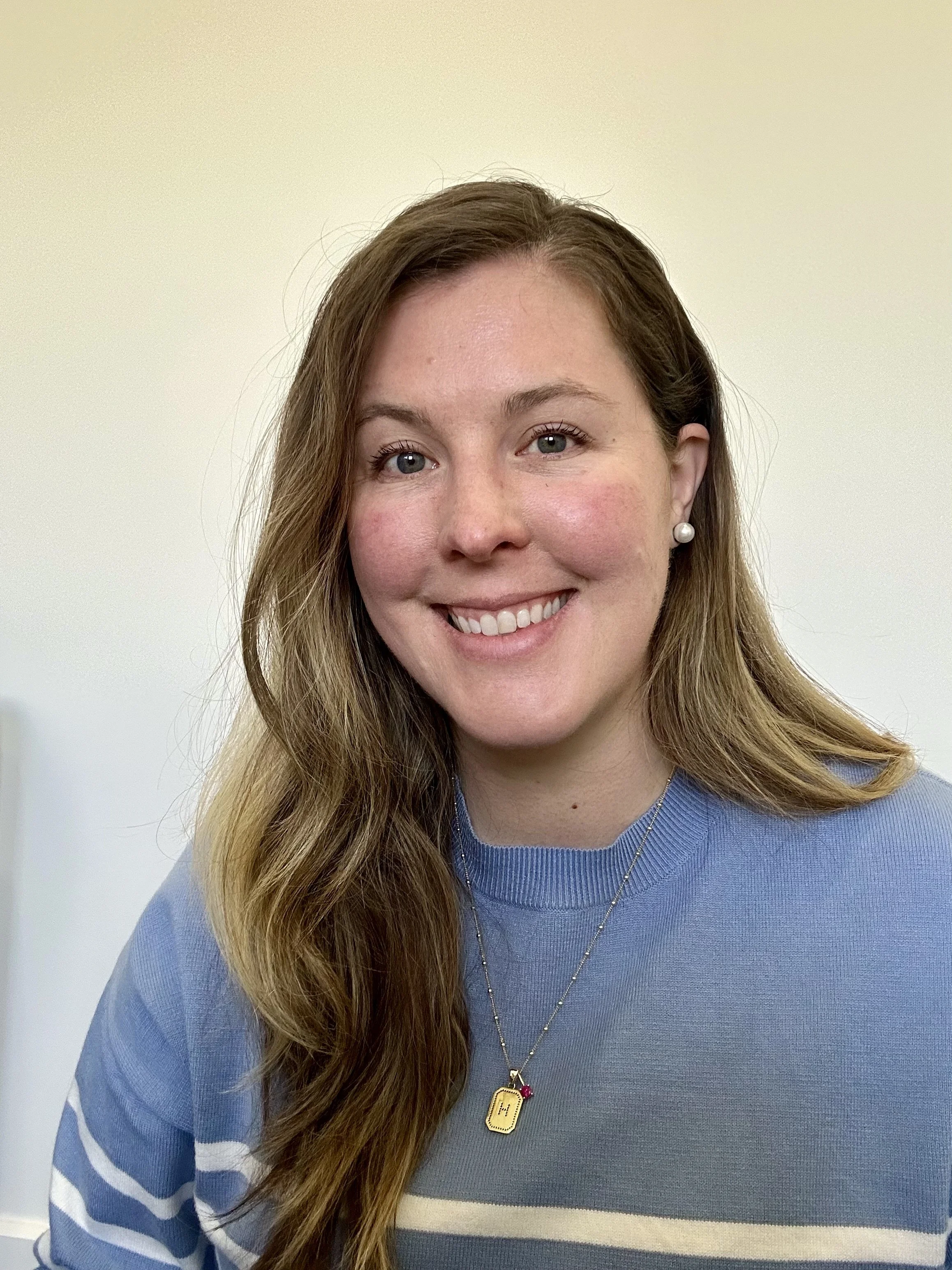 Close-up of a smiling woman with long wavy brown hair, light blue eyes, wearing pearl earrings, a blue sweater with white and navy stripes, and a gold dog tag necklace.