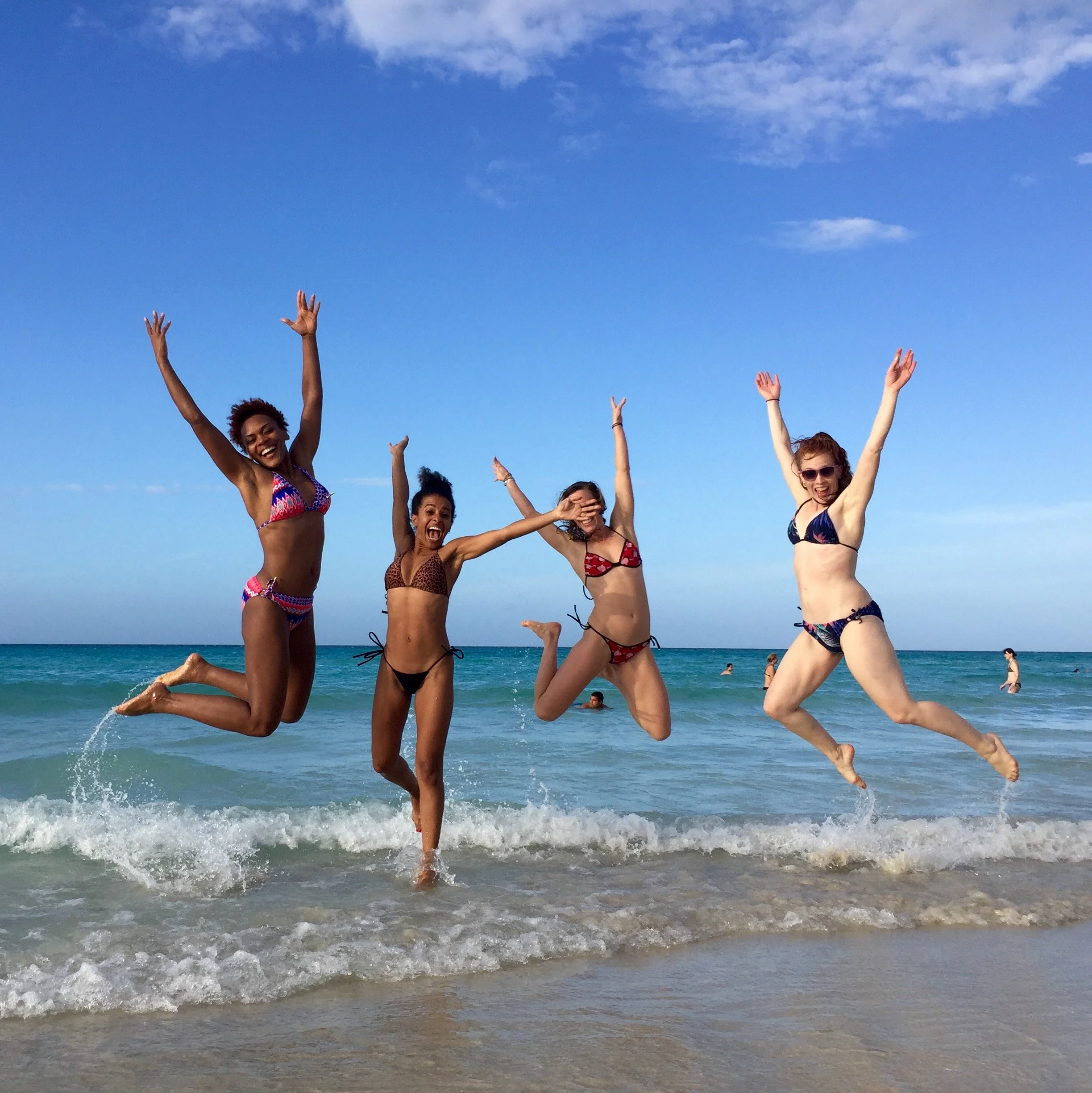 Group of women jumping and celebrating in the ocean on a sunny day, wearing swimsuits and sunglasses.