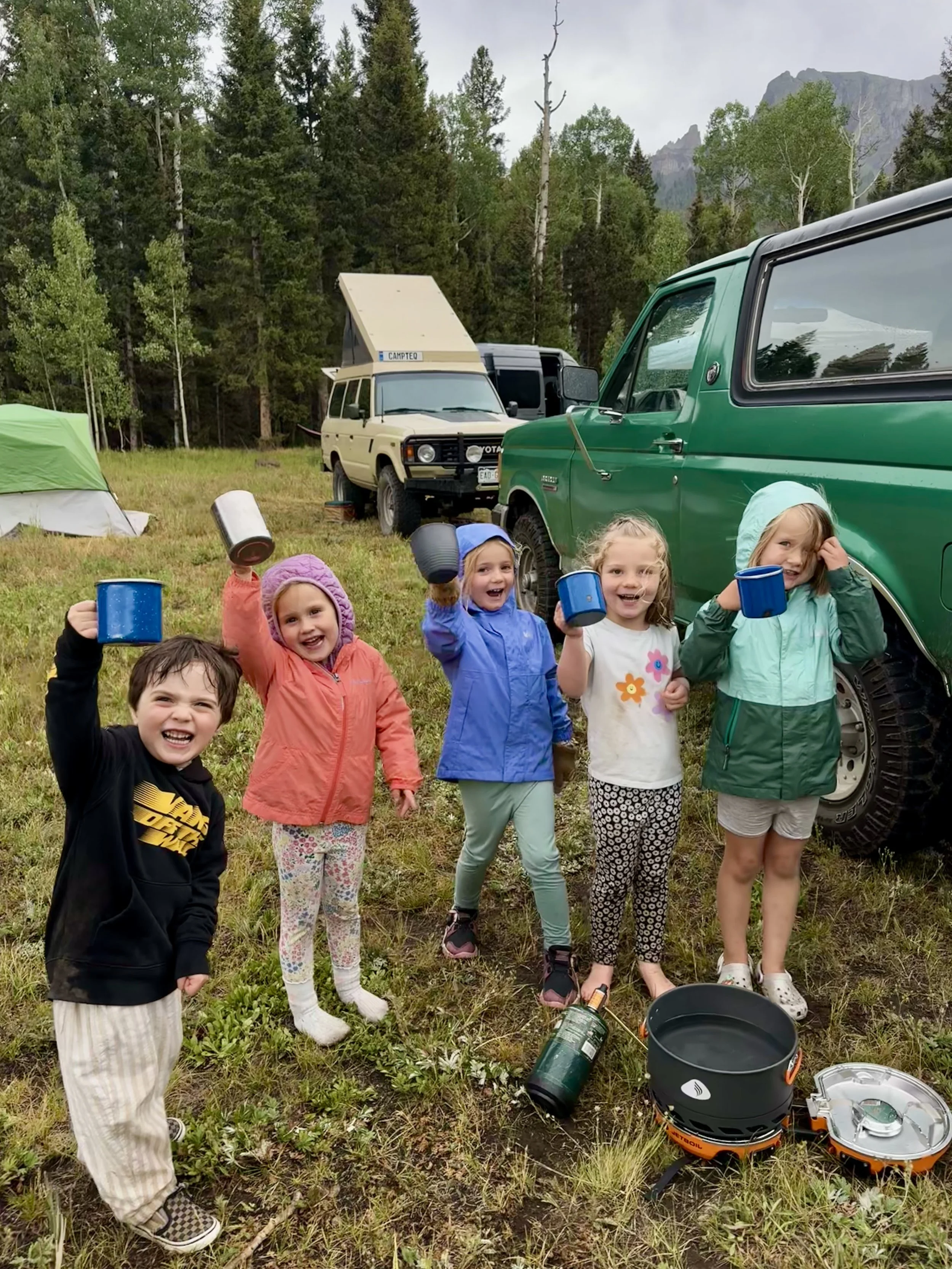A group of children camping in a grassy outdoor area with tents and vehicles, holding mugs and smiling, surrounded by trees and mountains in the background.