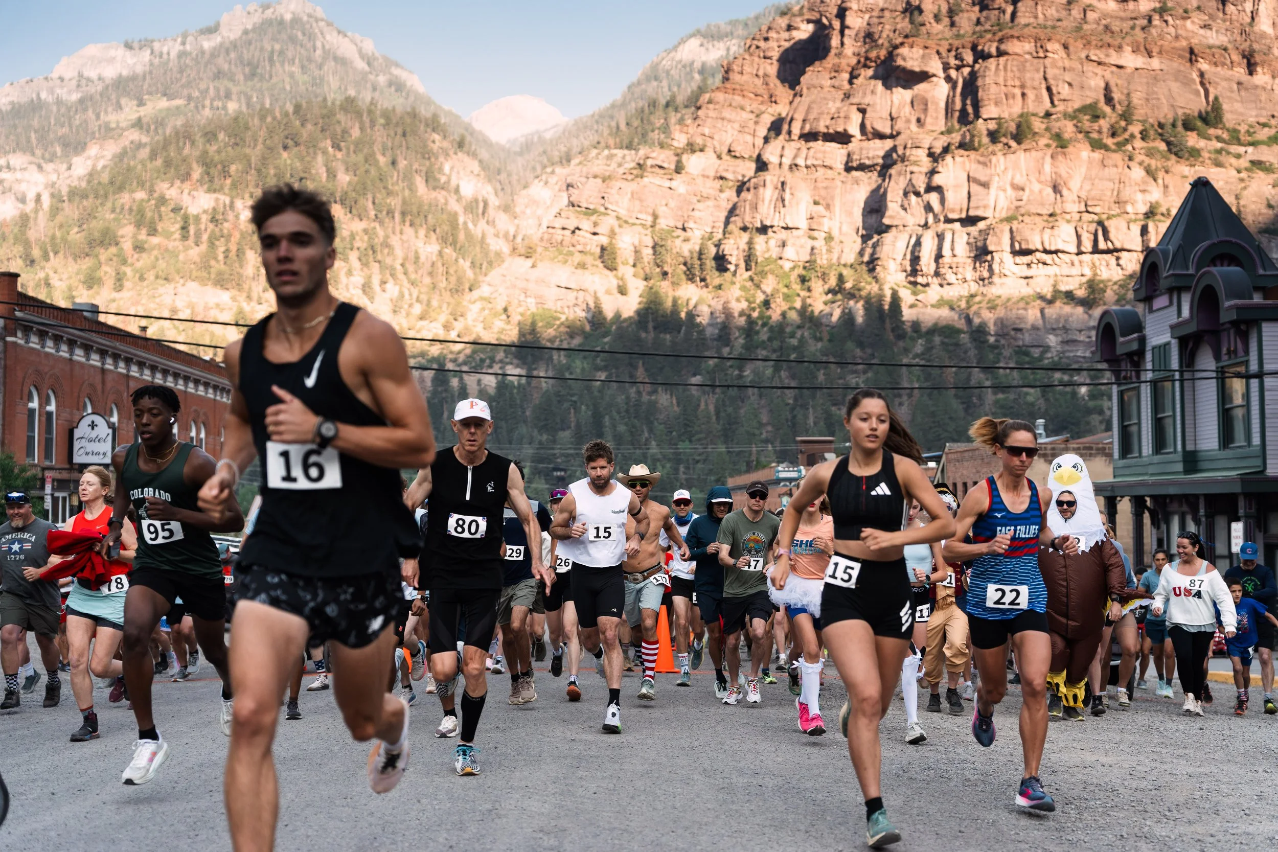 A group of runners participating in a race or marathon on a street with mountains in the background. The runners are wearing athletic clothing and numbered bibs, with some spectators visible along the street.