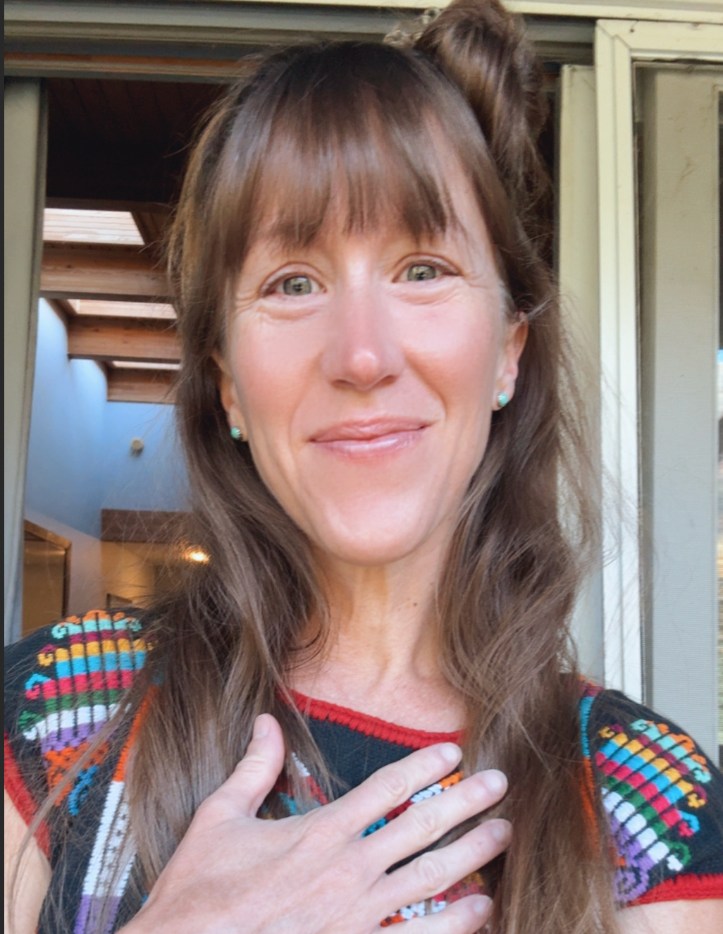 Close-up of a woman with long brown hair and green eyes, smiling and touching her chest, wearing a colorful, patterned top. Background shows a house or building with wooden ceiling beams and a skylight.