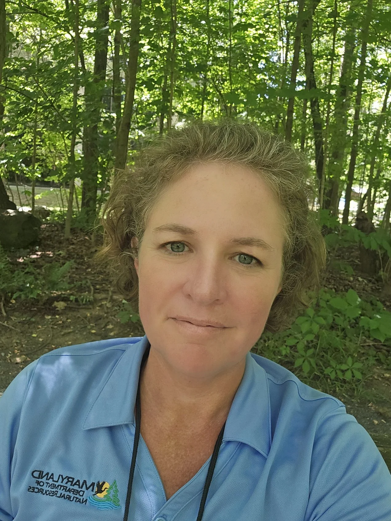 A woman with short curly hair and blue eyes taking a selfie outdoors in a wooded area, wearing a blue polo shirt with a logo for Madeira Department of Natural Resources and some trees in the background.