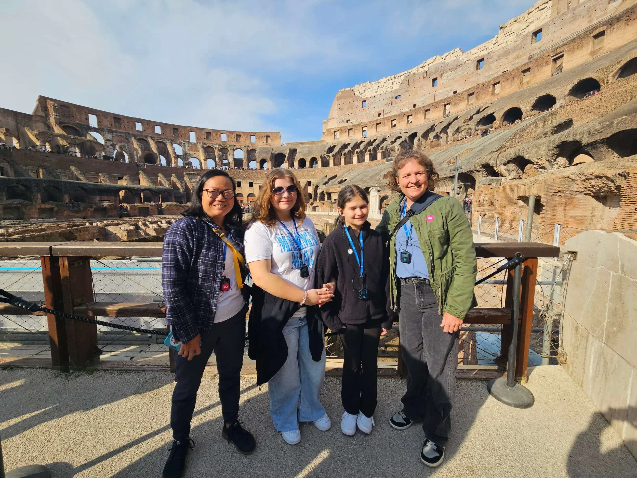 Four people stand smiling inside the Colosseum in Rome on a sunny day, with ancient stone arches in the background.