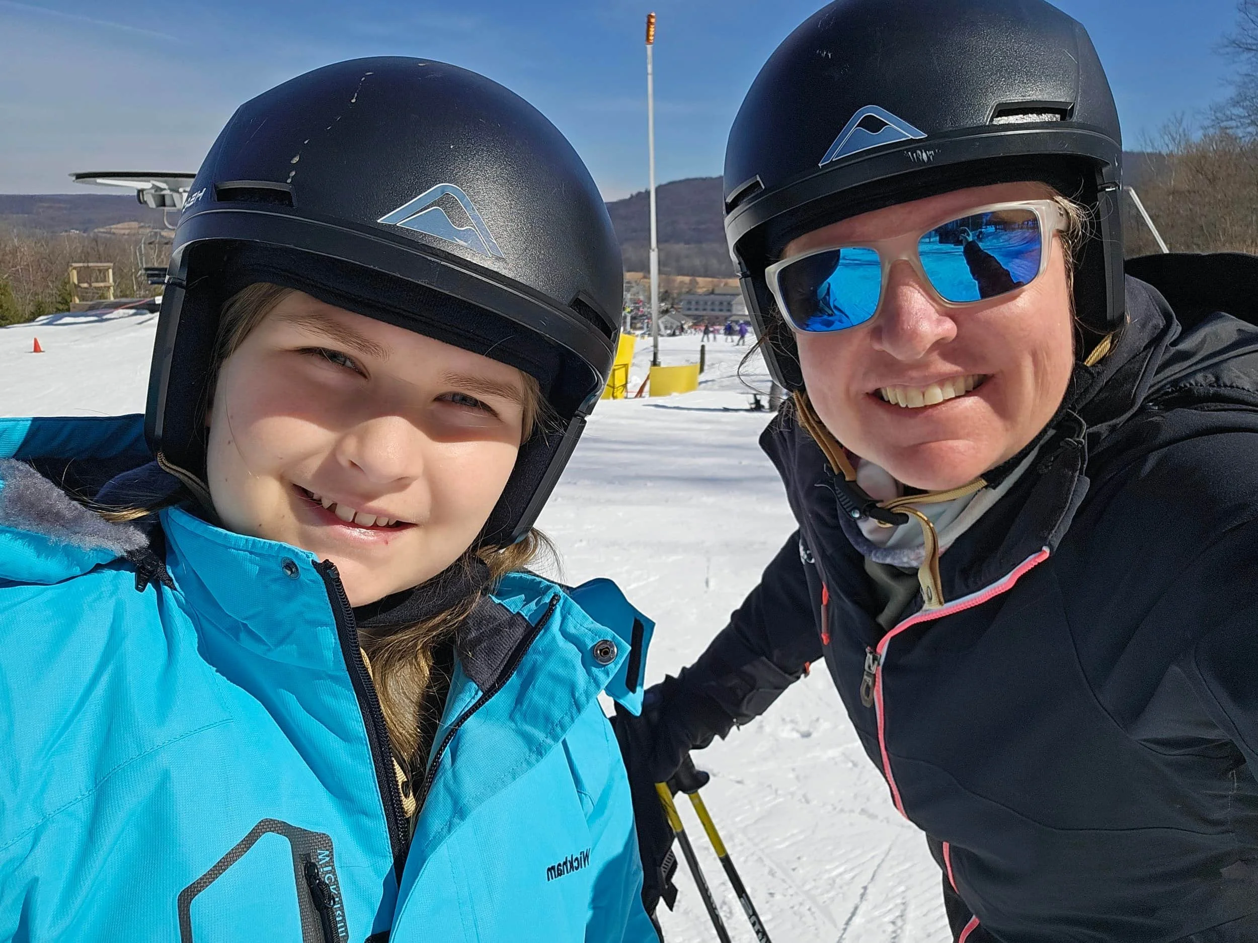 Two people in ski helmets and jackets smile on a sunny snowy slope with mountains and blue sky in the background.