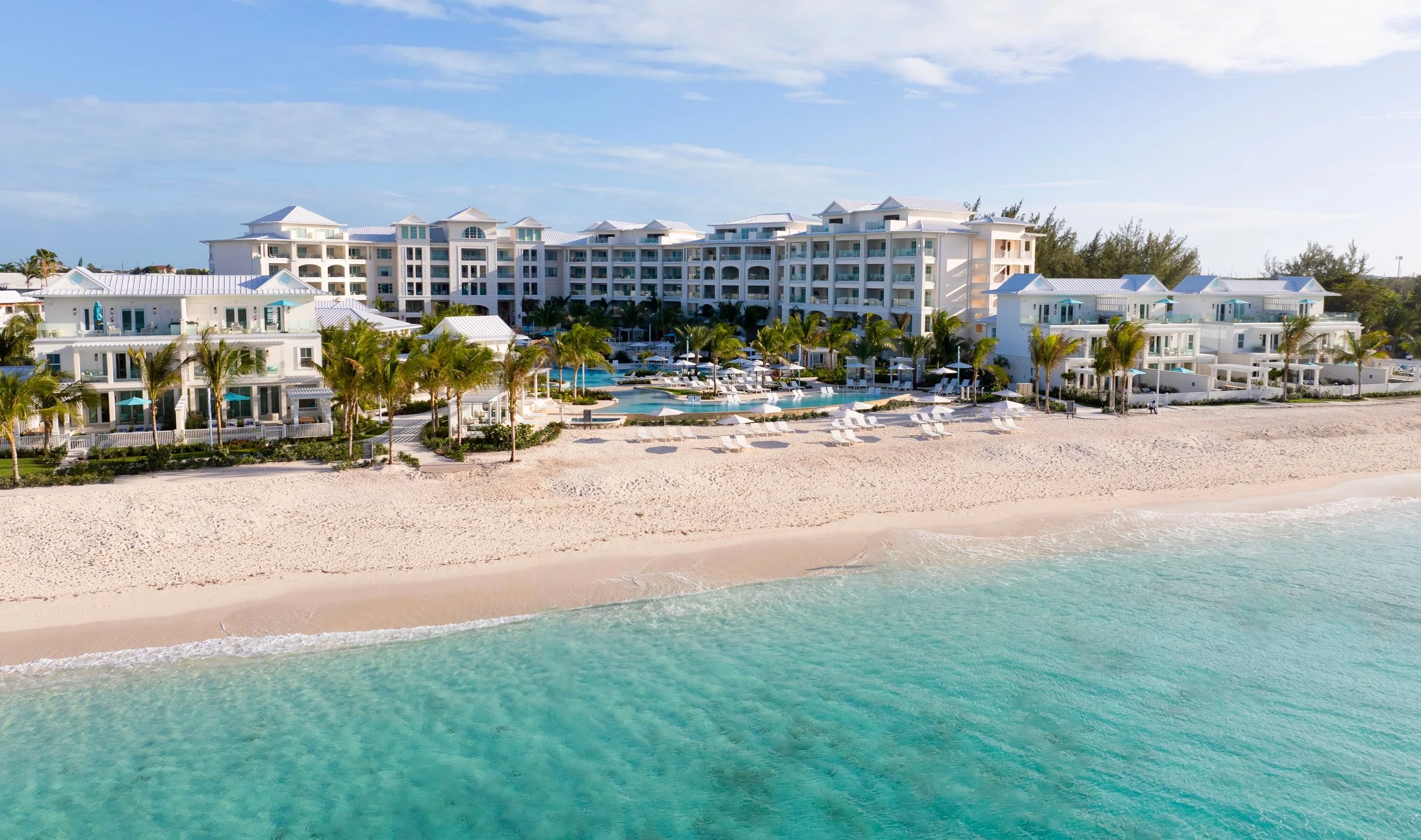 Beautiful beachfront resort with white buildings, palm trees, and a swimming pool, adjacent to a sandy beach and clear ocean water under a partly cloudy sky.