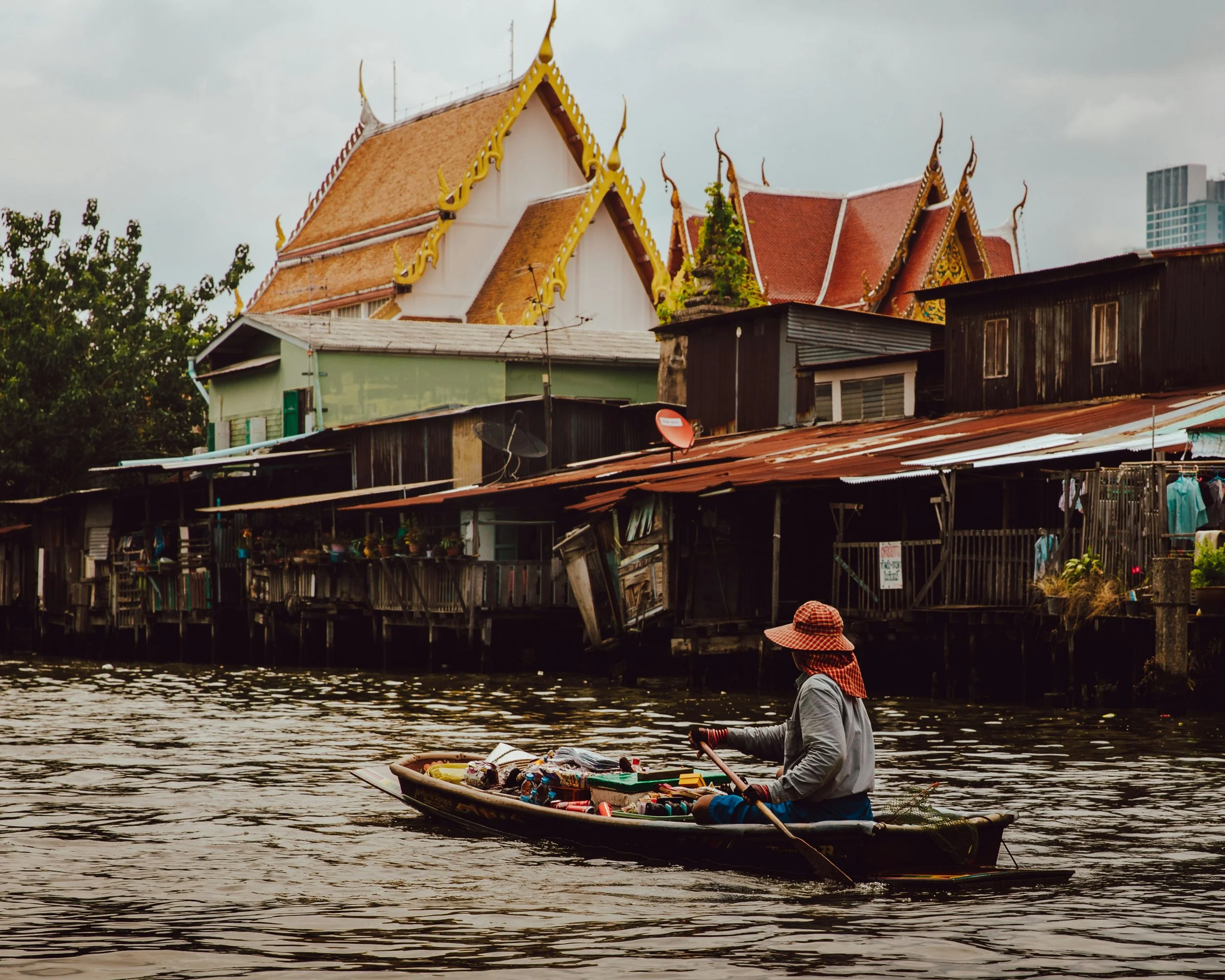 A person in a boat selling various goods along a river, with stilt houses and a traditional pointed roof temple in the background.
