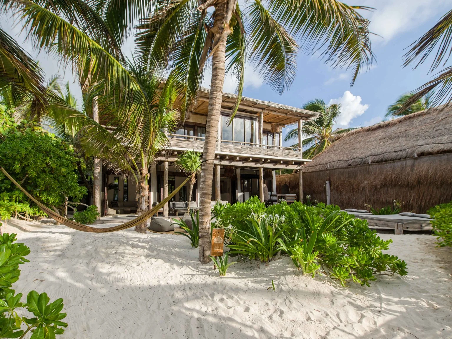 Beach house with wooden exterior surrounded by palm trees and green tropical plants, sandy beach, and lounge chairs.
