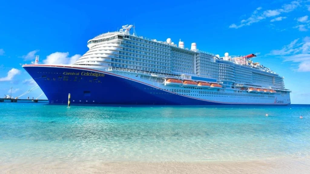 A large cruise ship docked in clear blue waters near a sandy beach under a partly cloudy sky.