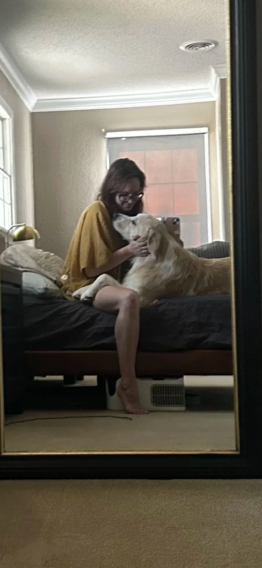 A woman sitting on a bed, taking a mirror selfie with a dog snuggling against her, in a bedroom with beige walls and a window in the background.