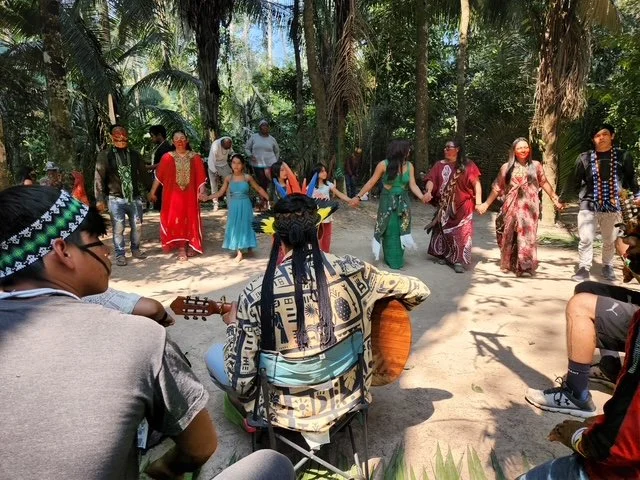 Group of people holding hands in a circle, dancing outdoors in a tropical setting with tall trees and sunlight.