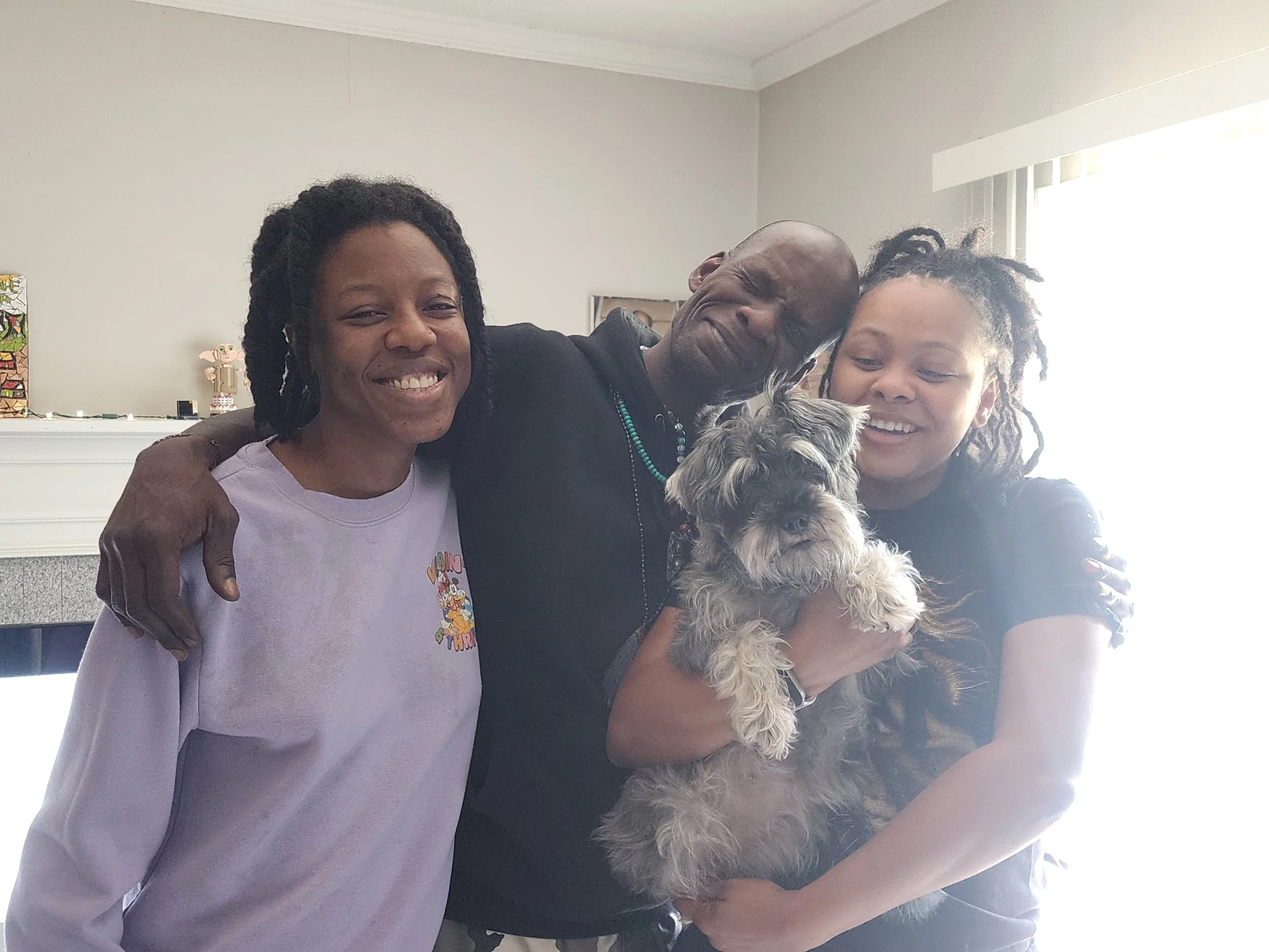 Three people smiling and hugging, holding a small dog indoors near a window with blinds.