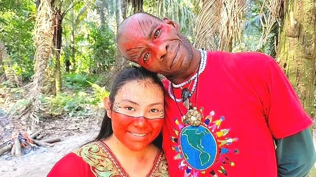 A man and a woman smiling outdoors in a forest, with red face paint or markings, wearing colorful clothing and accessories.