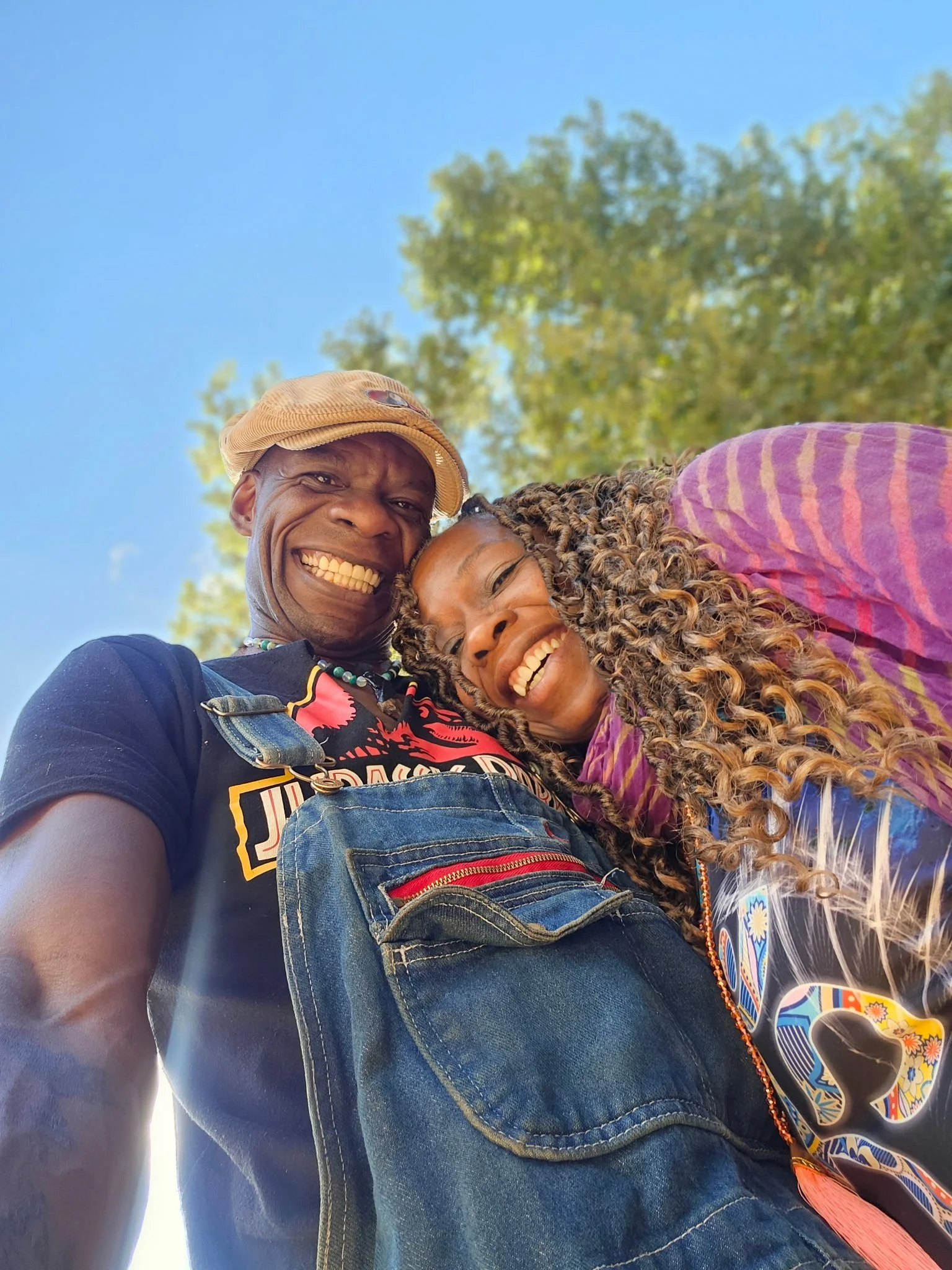A smiling man and woman taking a selfie outdoors, with a clear blue sky and trees in the background.
