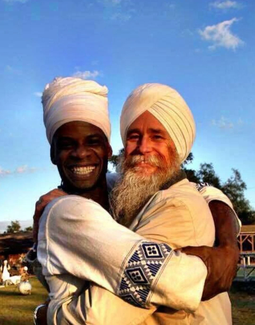 Two men wearing traditional Indian turbans hugging and smiling outdoors under a blue sky.