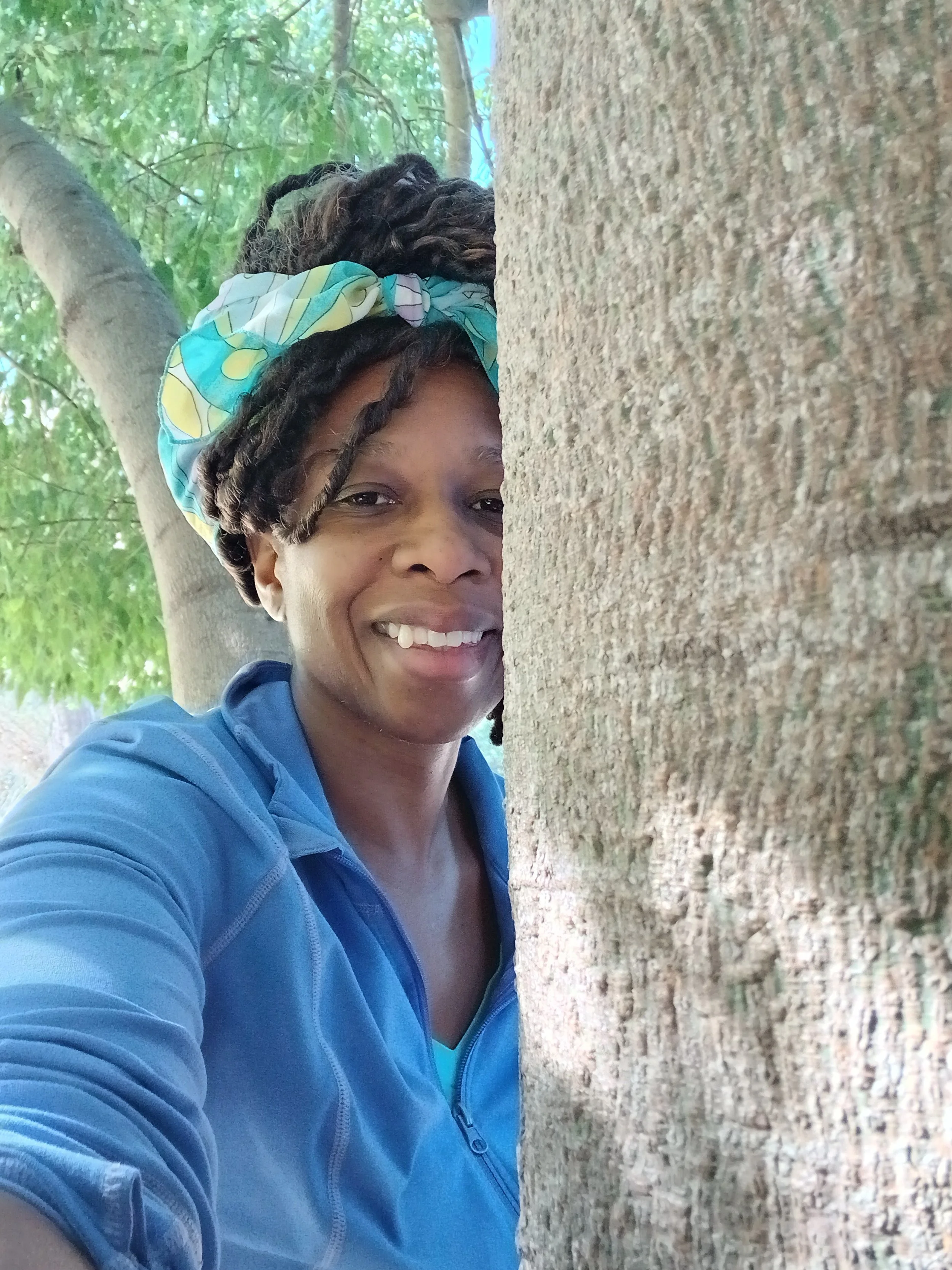 A woman smiling and leaning on a large tree trunk outdoors during daytime, with green leaves and branches visible in the background, wearing a blue jacket and a colorful head scarf.