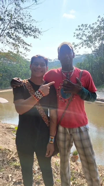 Two men standing outdoors near a body of water with trees and sky in the background. One man is smiling and pointing at the other, who is wearing a red shirt and making a peace sign with his hand.