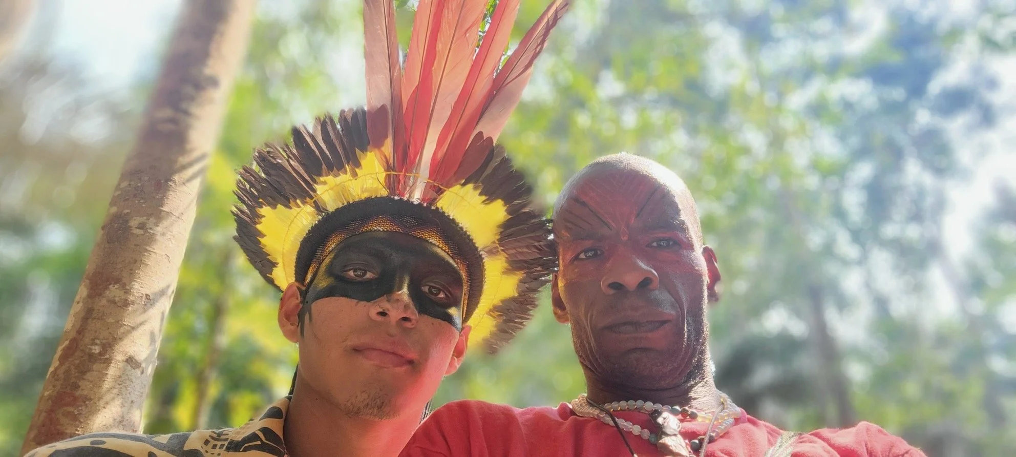 Two men taking a selfie outdoors, with one wearing traditional indigenous headdress and face paint, and the other wearing red clothing and jewelry, trees and sunlight in the background.