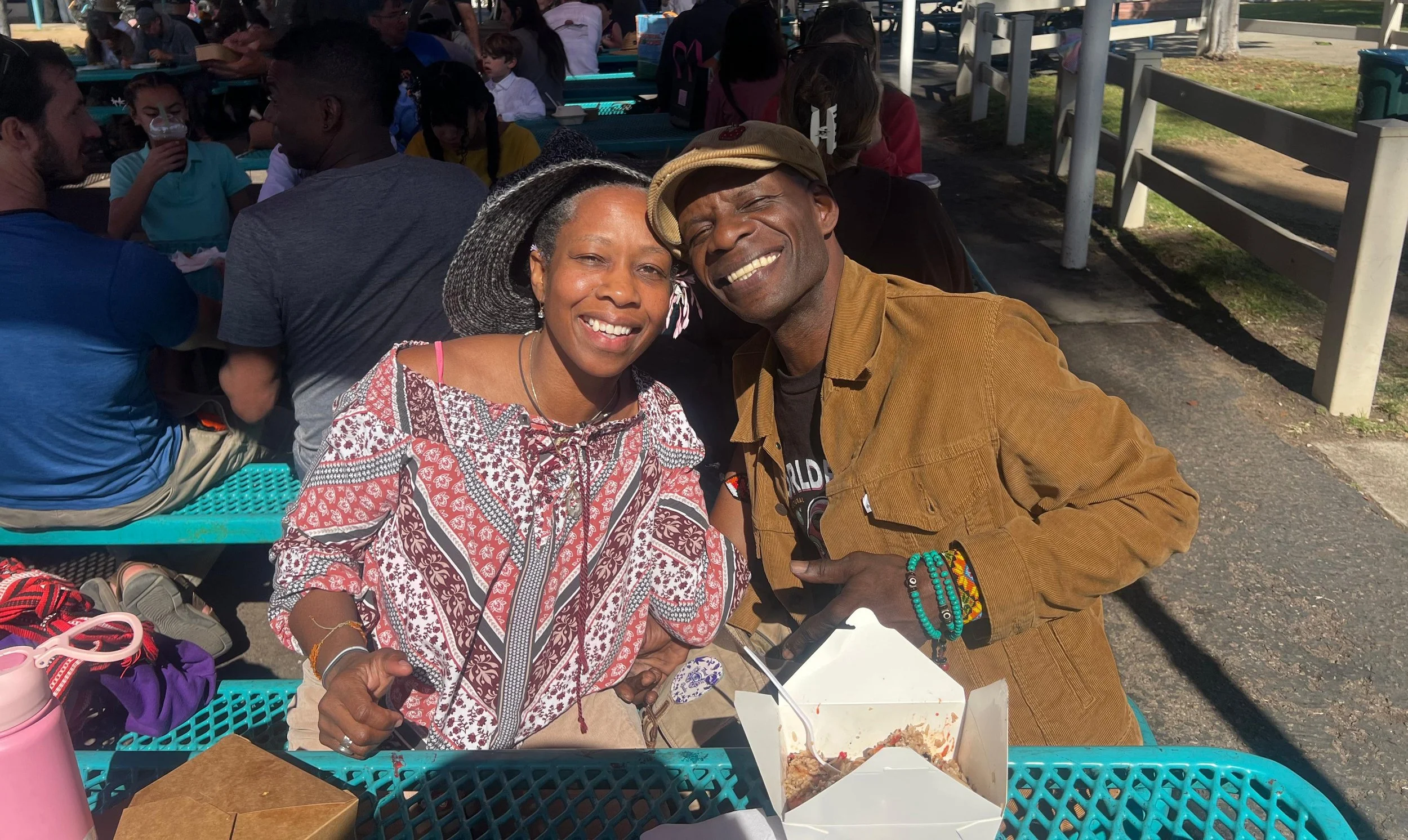 Two people smiling and sitting at a picnic table outdoors, with other people in the background at a crowded gathering or event.