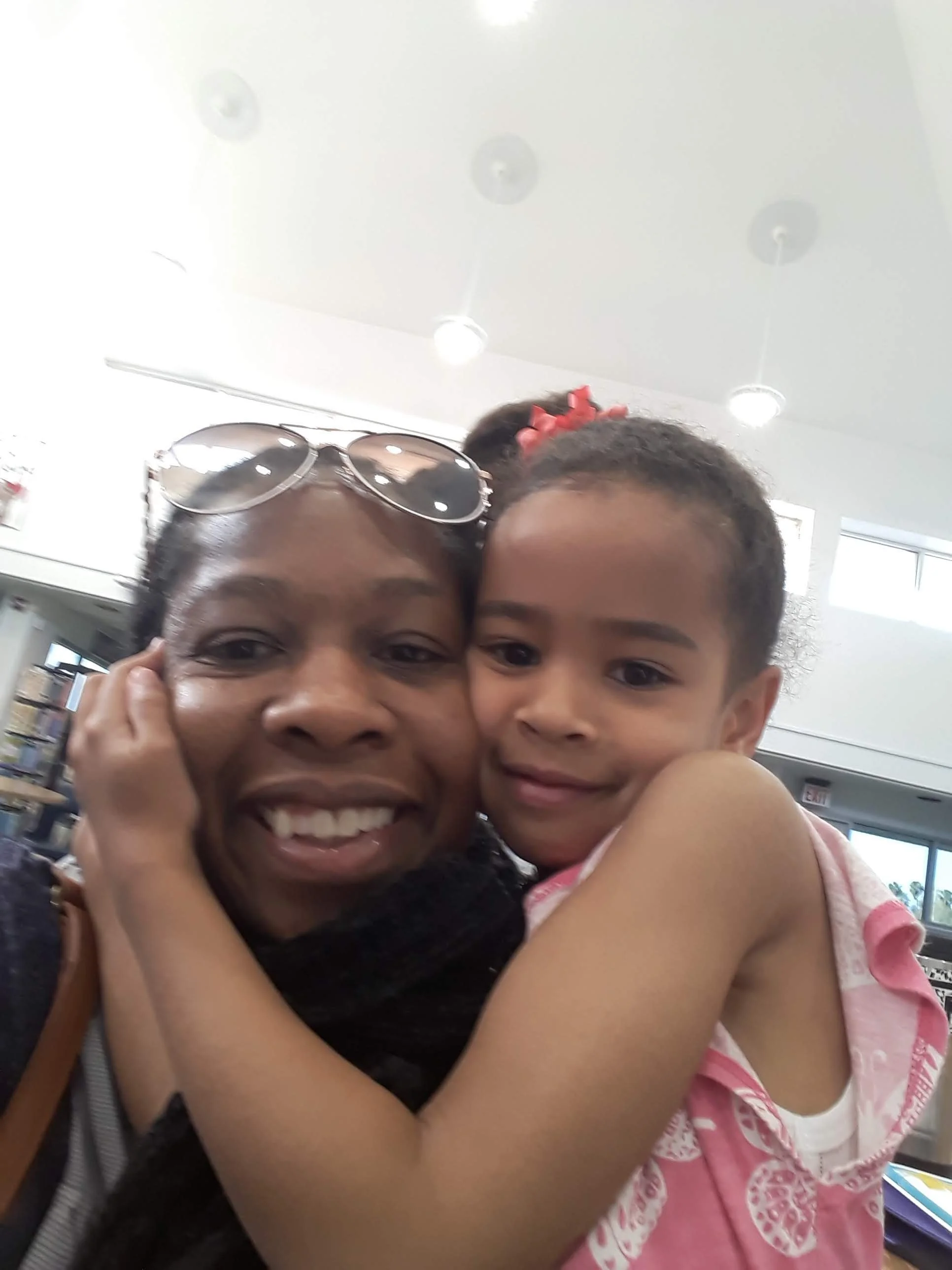 A woman and a young girl are hugging and smiling at the camera. The woman is wearing glasses on her head, and the girl has a pink bow in her hair.
