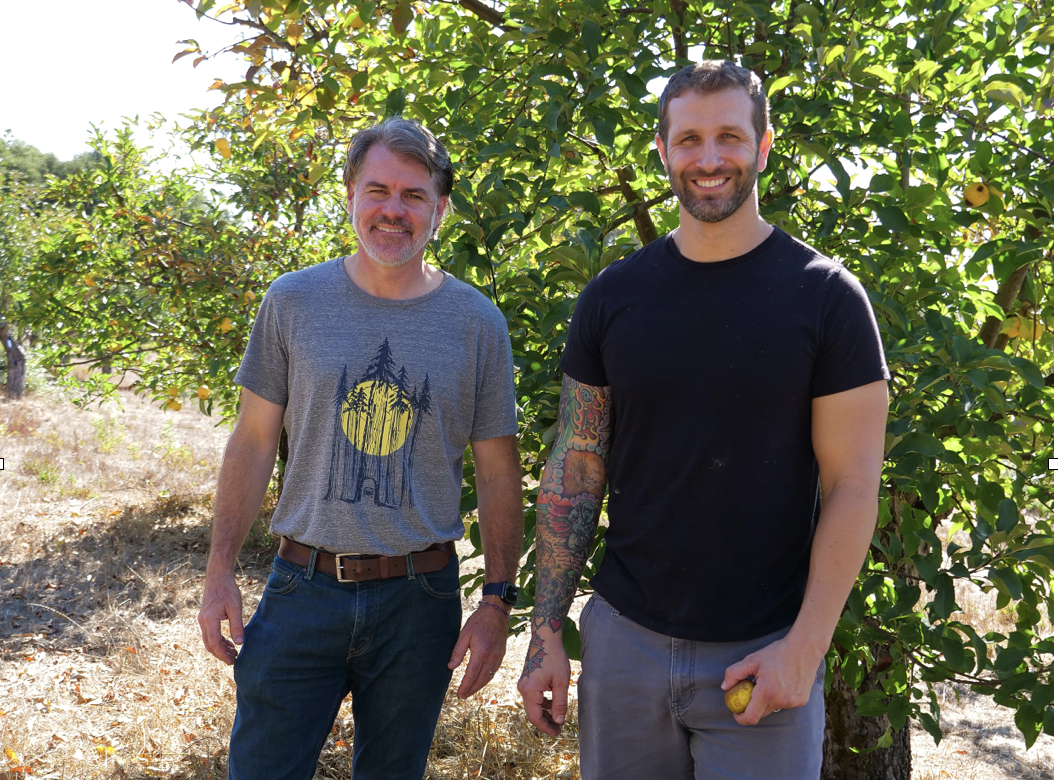 Two men standing outdoors among lush green trees, smiling at the camera, with sunlight filtering through the leaves.