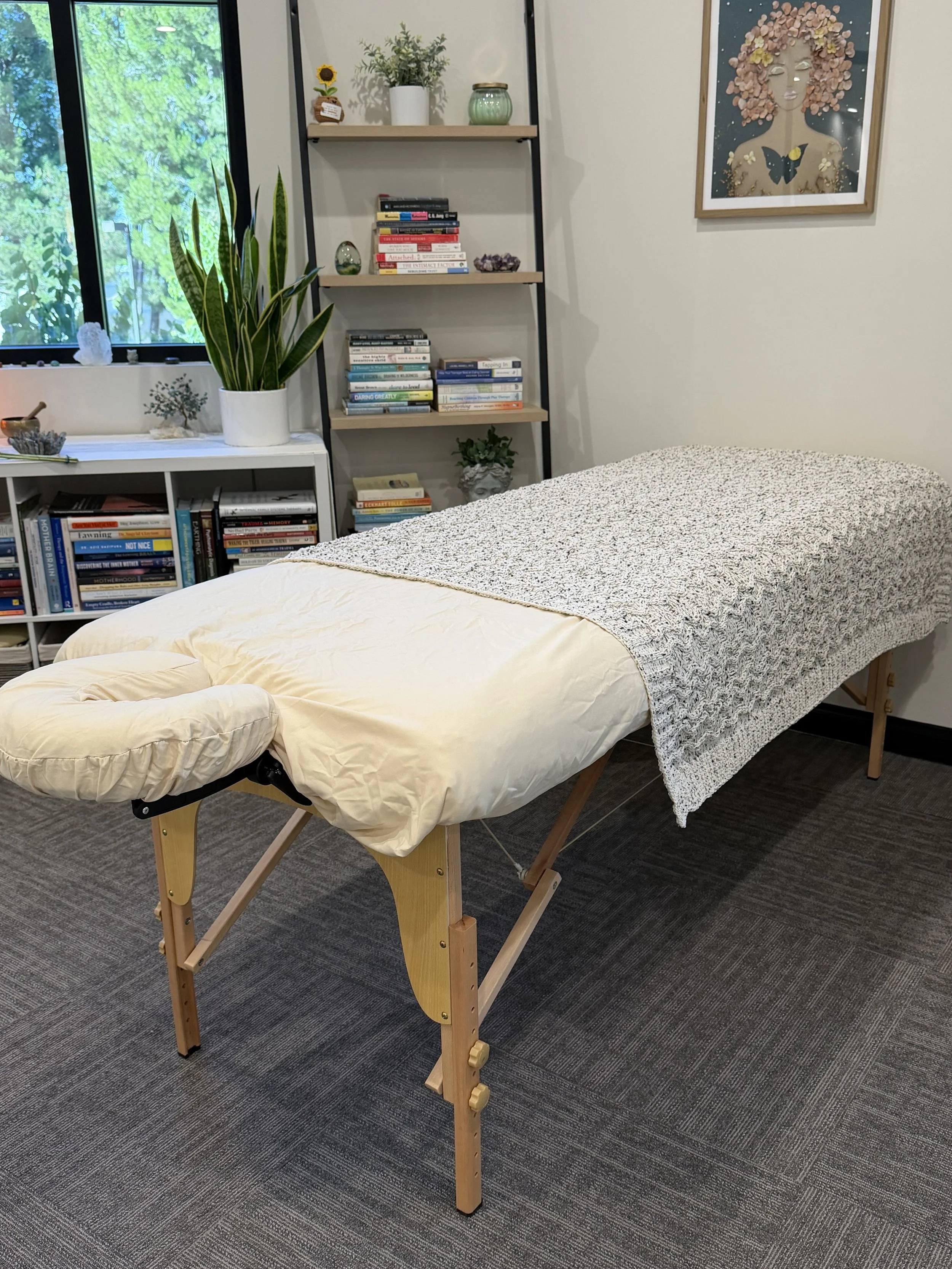 A massage table with a cream-colored sheet and a textured gray blanket in a cozy room. The room has bookshelf with books and plants, a window with green trees outside, a framed artwork of a woman with purple hair, and decorative items including a sunflower plant and a container with salt crystals.